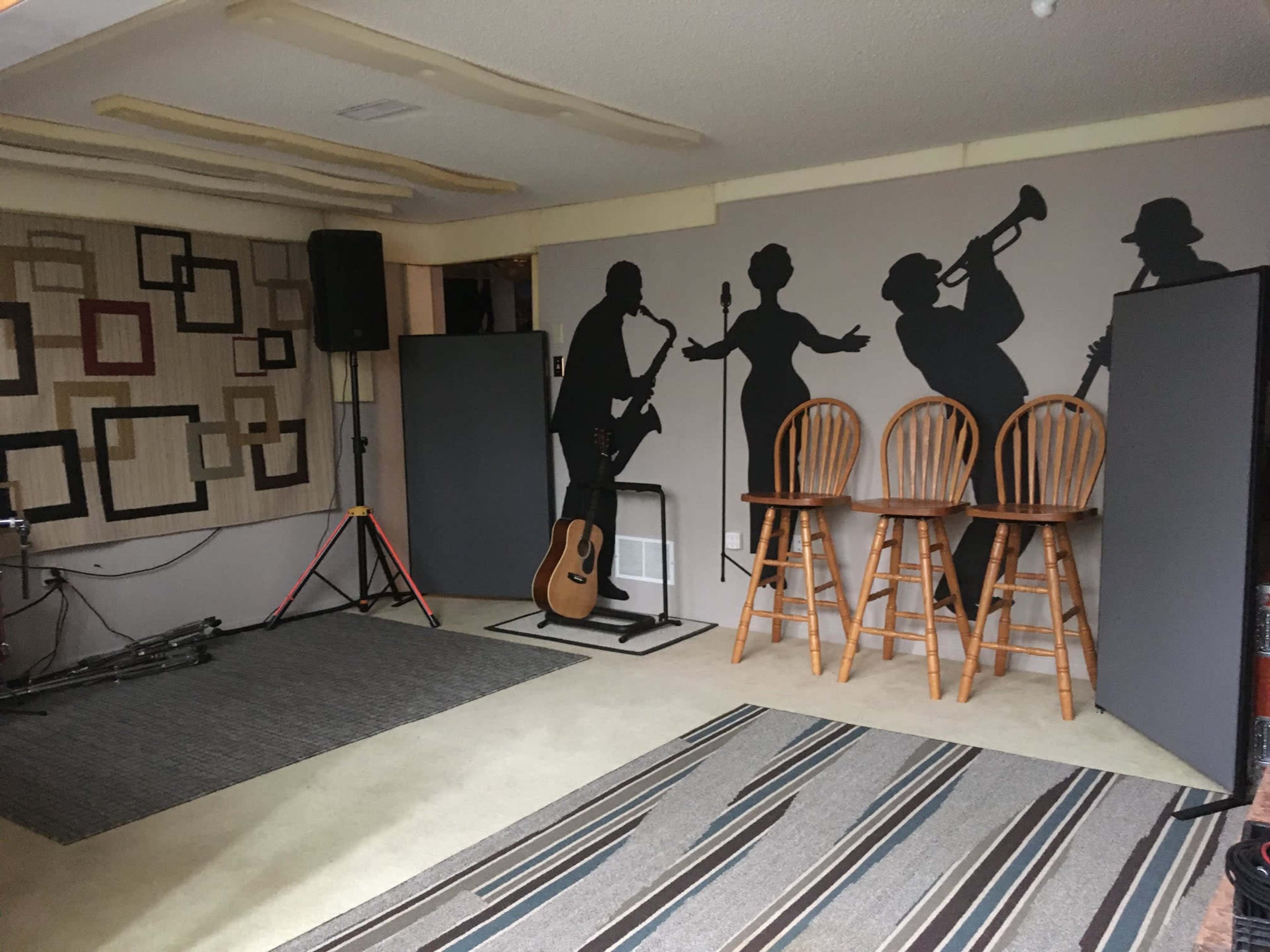 The image shows a music practice room featuring silhouettes of musicians on the walls, three wooden stools, an acoustic guitar, and a sound system setup.
