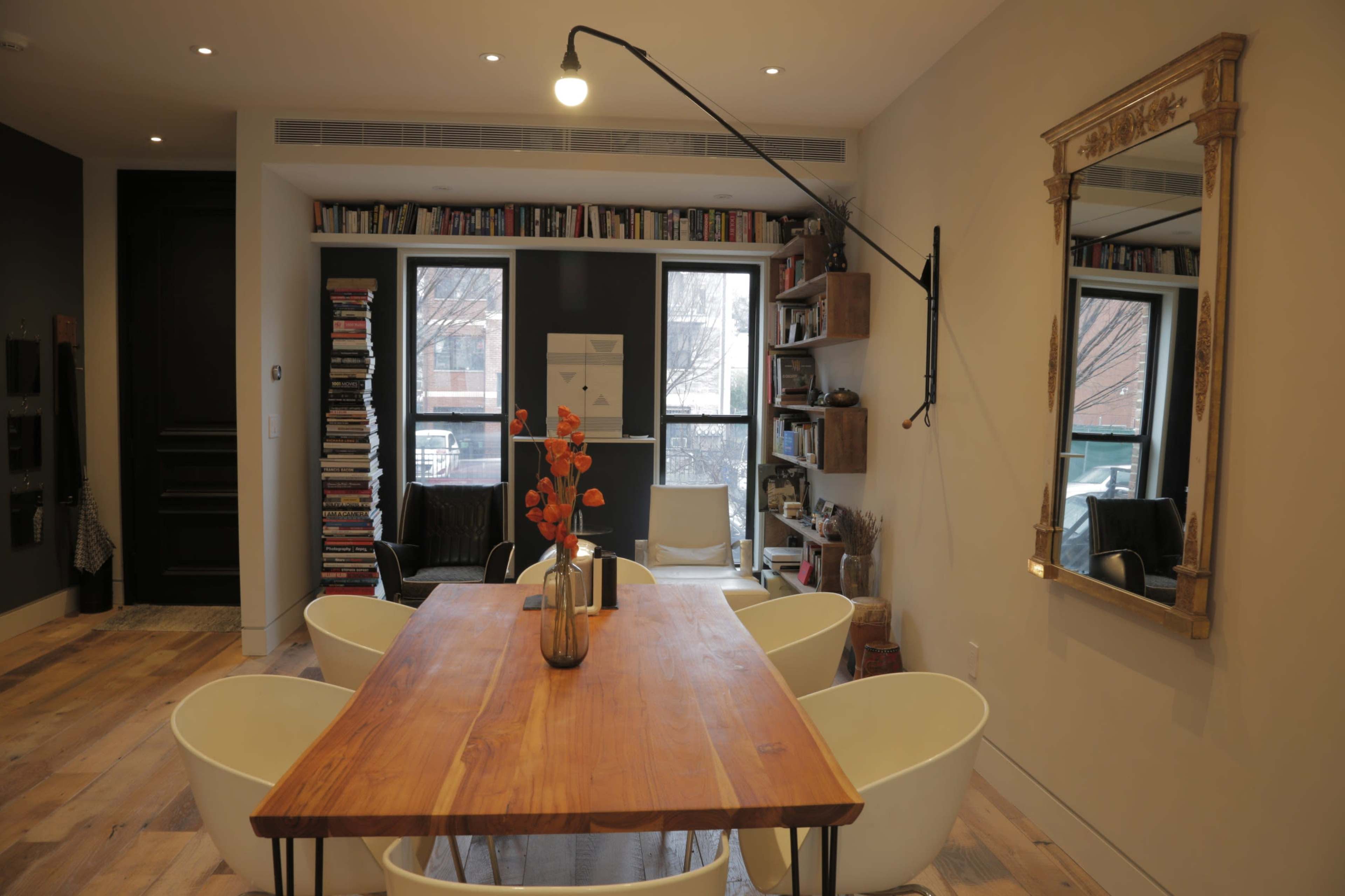 A dining area features a wooden table surrounded by four white chairs, with bookshelves and large windows in the background.
