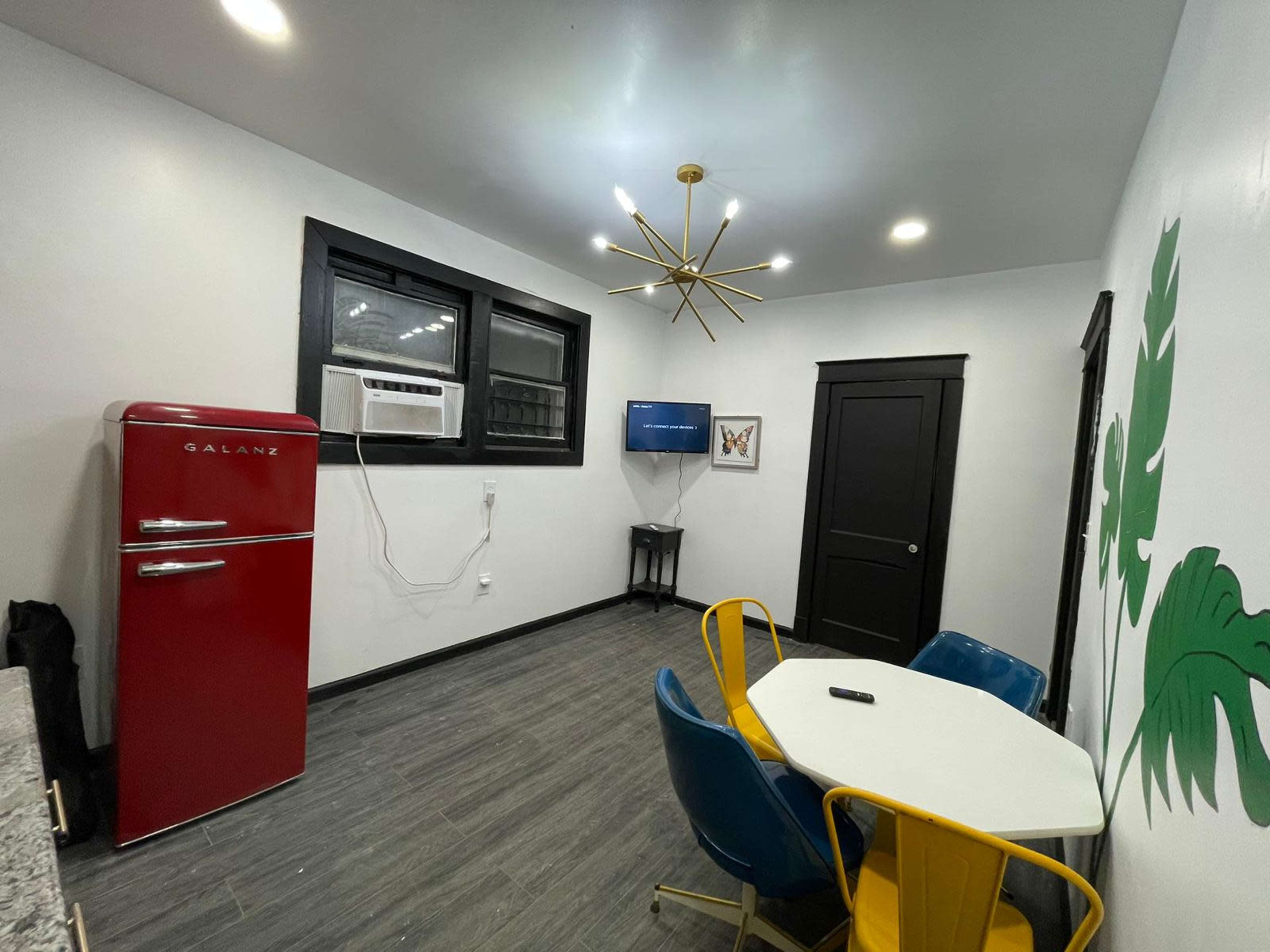 The image features a modern kitchen area with a red retro-style refrigerator, an air conditioning unit, a small round table surrounded by blue and yellow chairs, and a wall with a green leaf design.