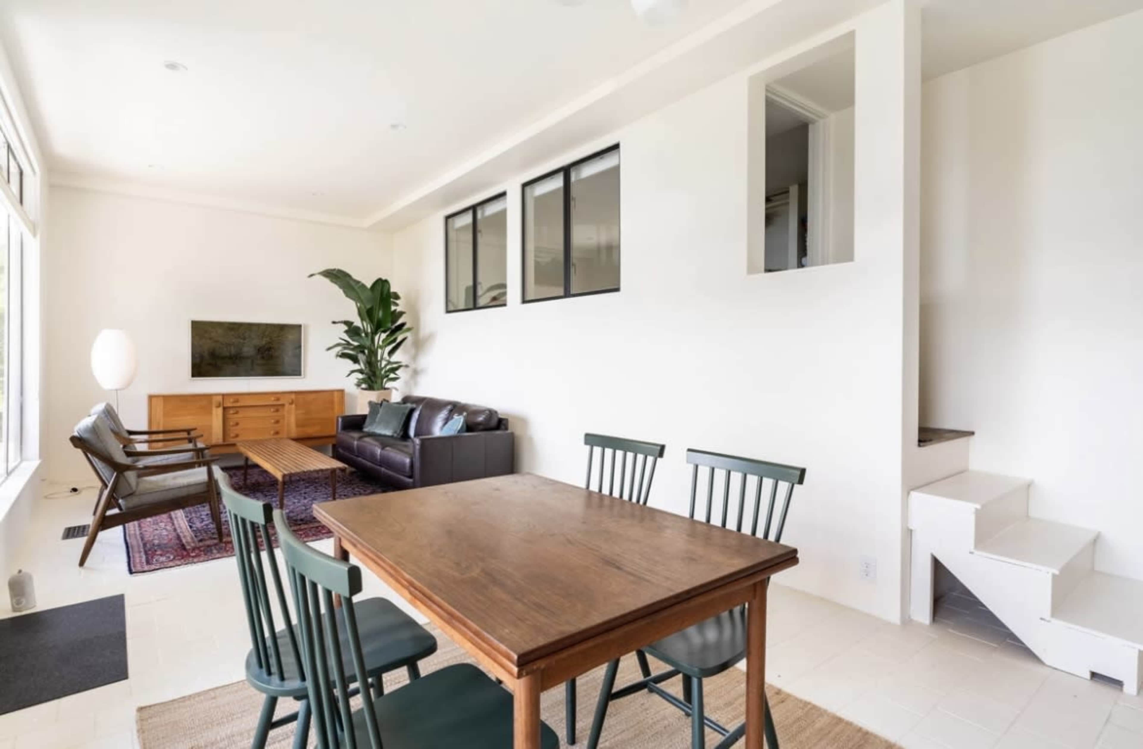 A spacious living area features a dining table, a leather sofa, an entertainment unit, and large windows that allow natural light to enter.