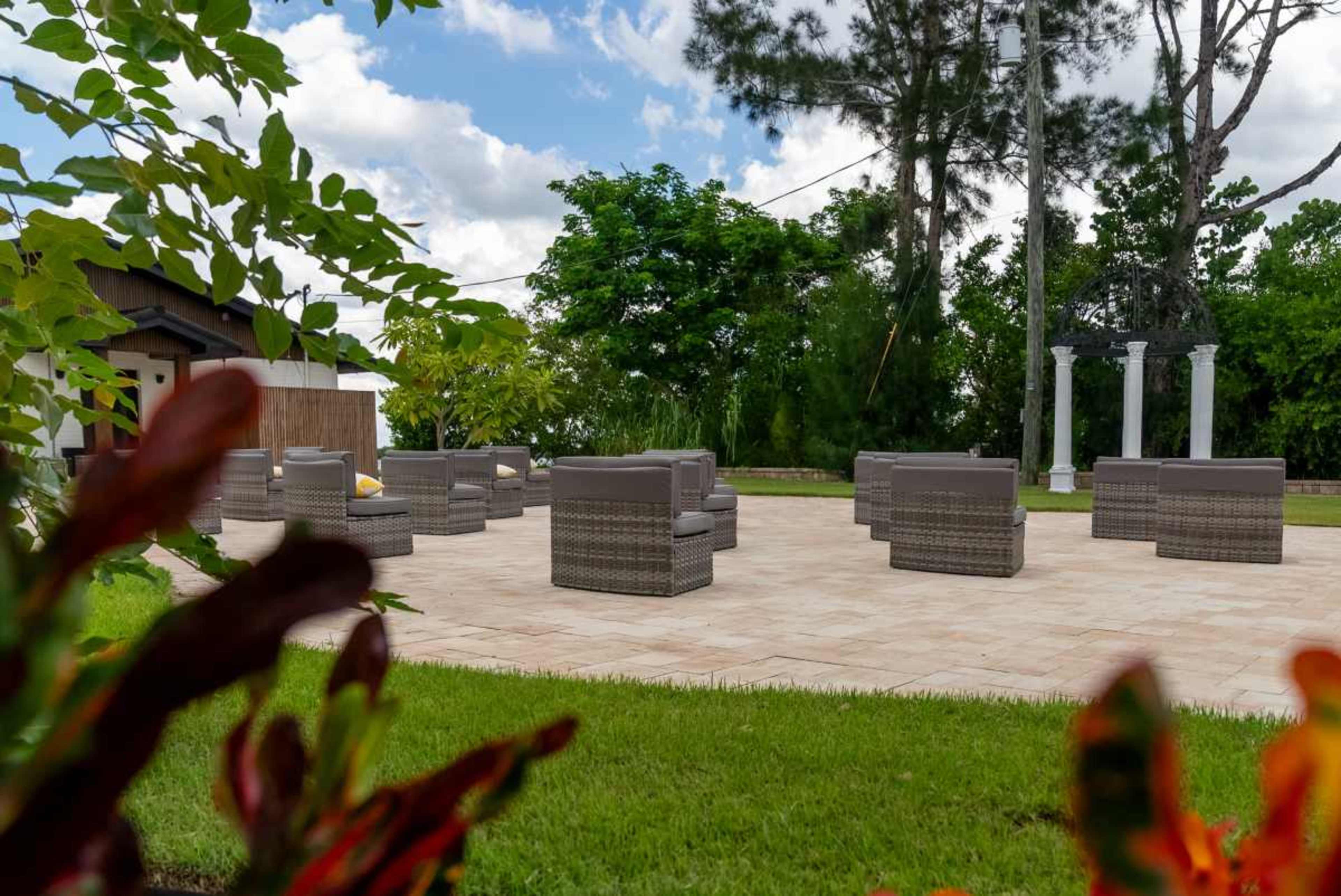 A landscaped outdoor area features a stone patio with several wicker seating arrangements and decorative foliage in the foreground.