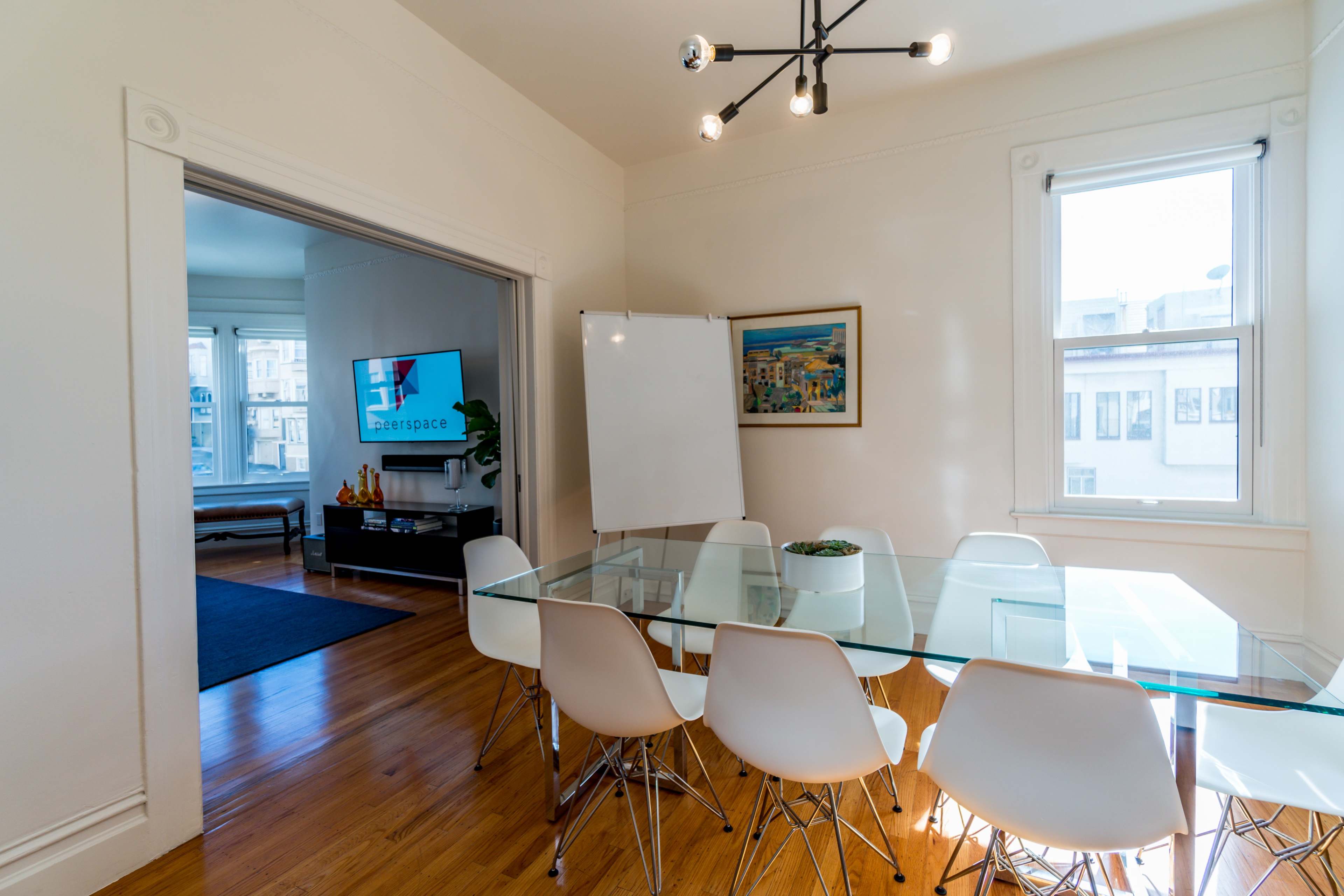 A glass dining table surrounded by white chairs is positioned in a bright room, with a TV and a whiteboard visible in an adjoining space.