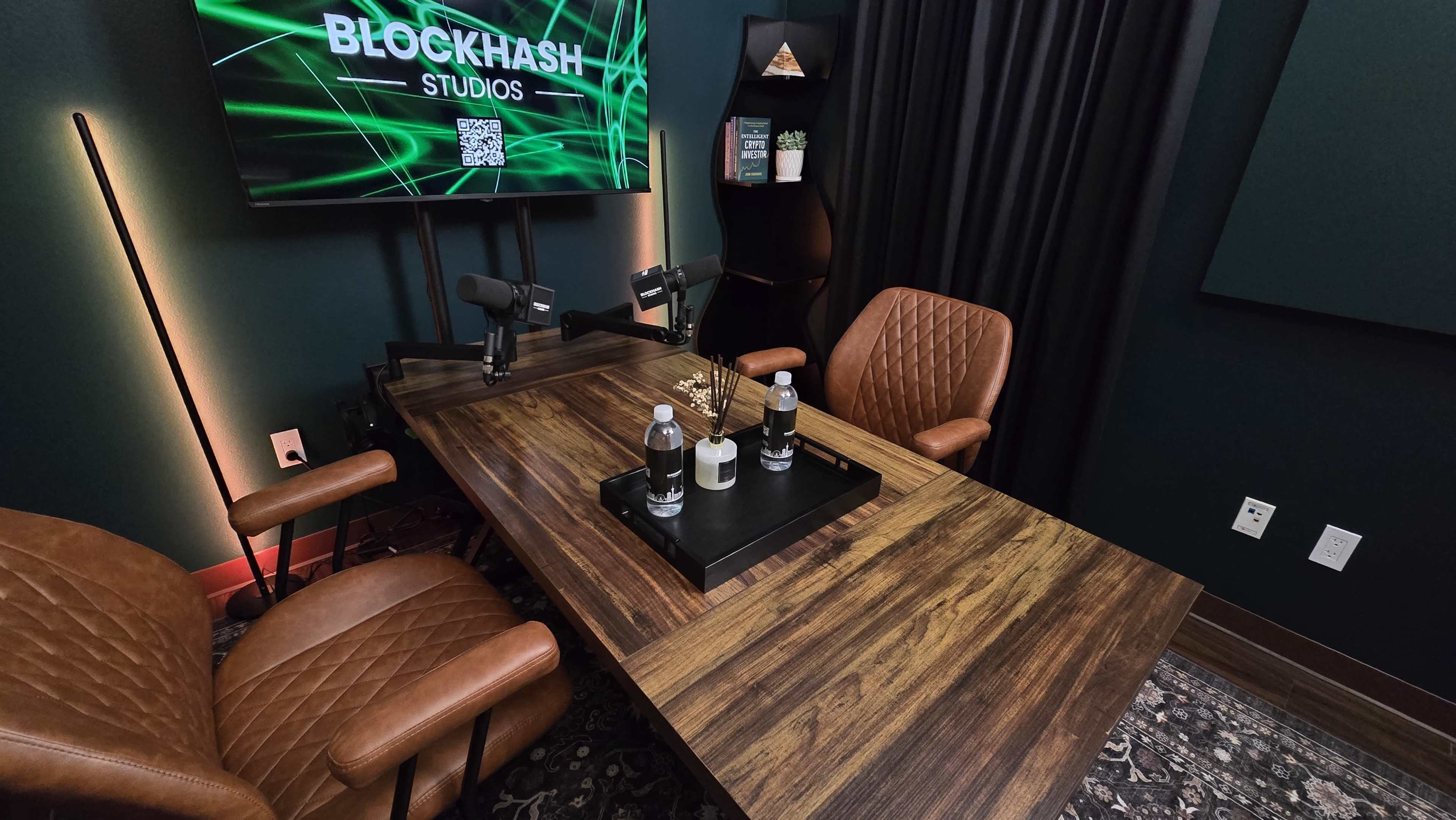 The image shows a podcast studio setup featuring a wooden table with two microphones, two chairs, and bottles of water, alongside a backdrop with green digital graphics and a display screen labeled "BLOCKHASH STUDIOS."