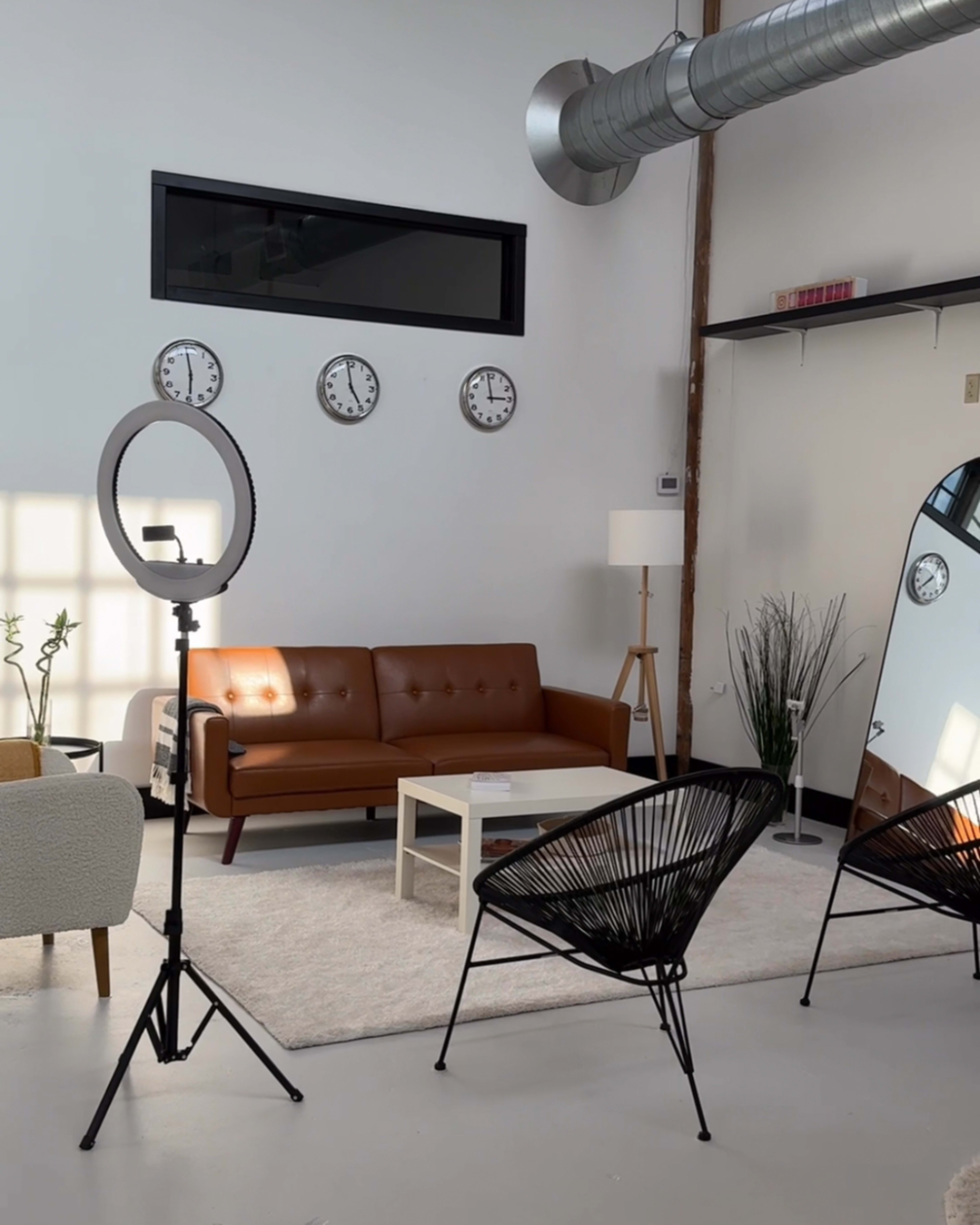 A modern interior space featuring a brown leather sofa, a white coffee table, three wall clocks, and two black chairs beside a large mirror.
