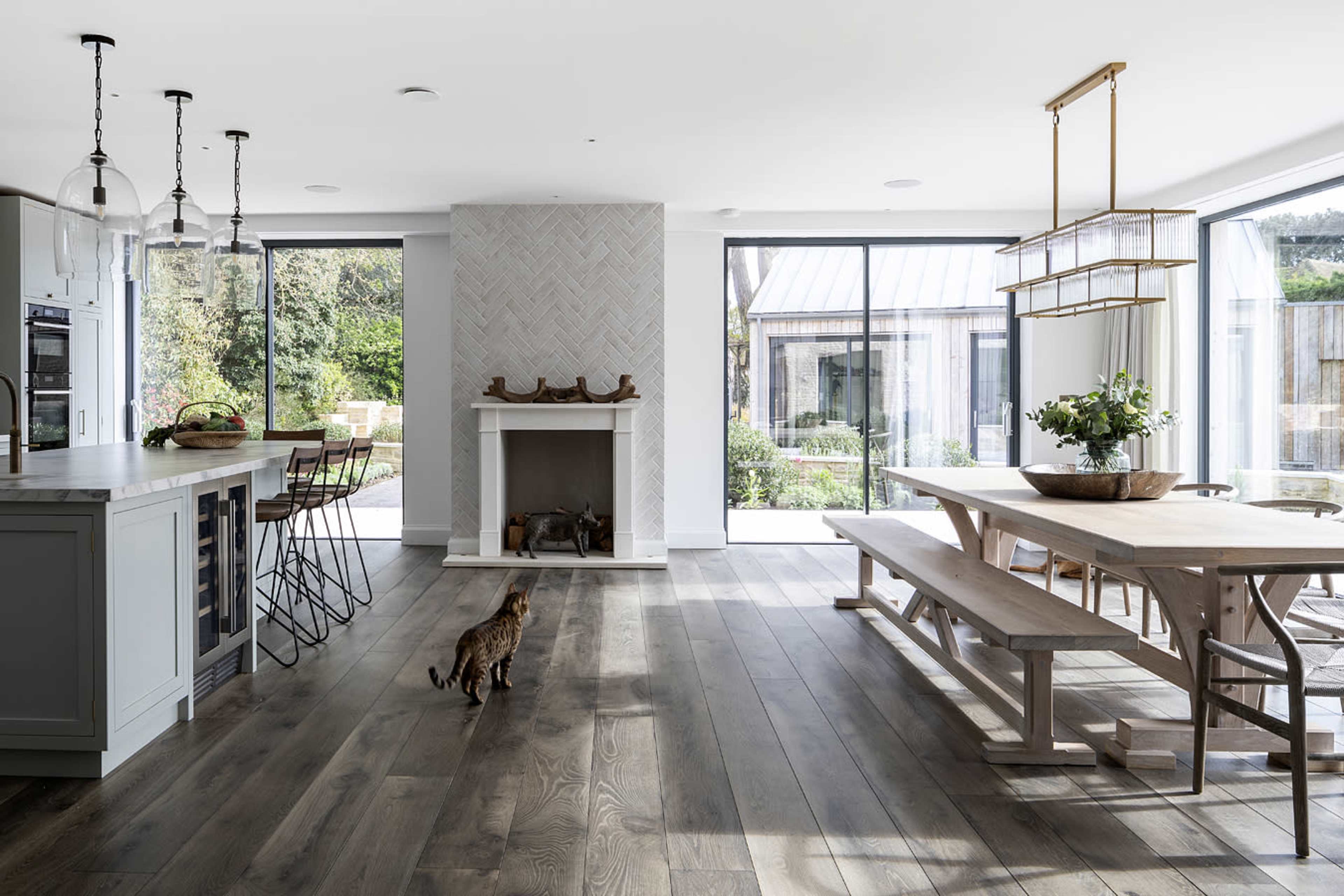 A modern kitchen and dining area features a large wooden table, a central island with bar stools, a mantelpiece, and large windows overlooking a garden.