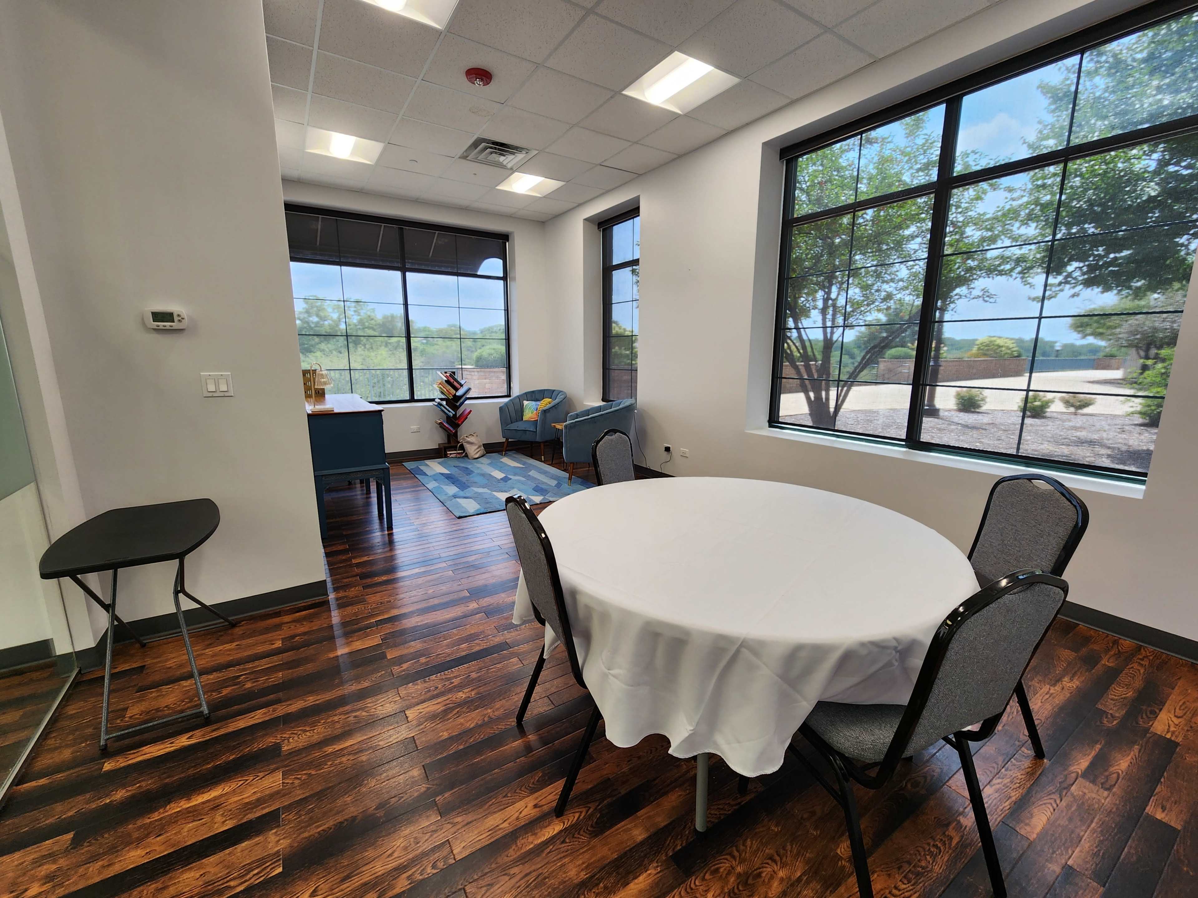 A well-lit meeting room featuring a round table with chairs, large windows overlooking a landscape, and a small seating area.