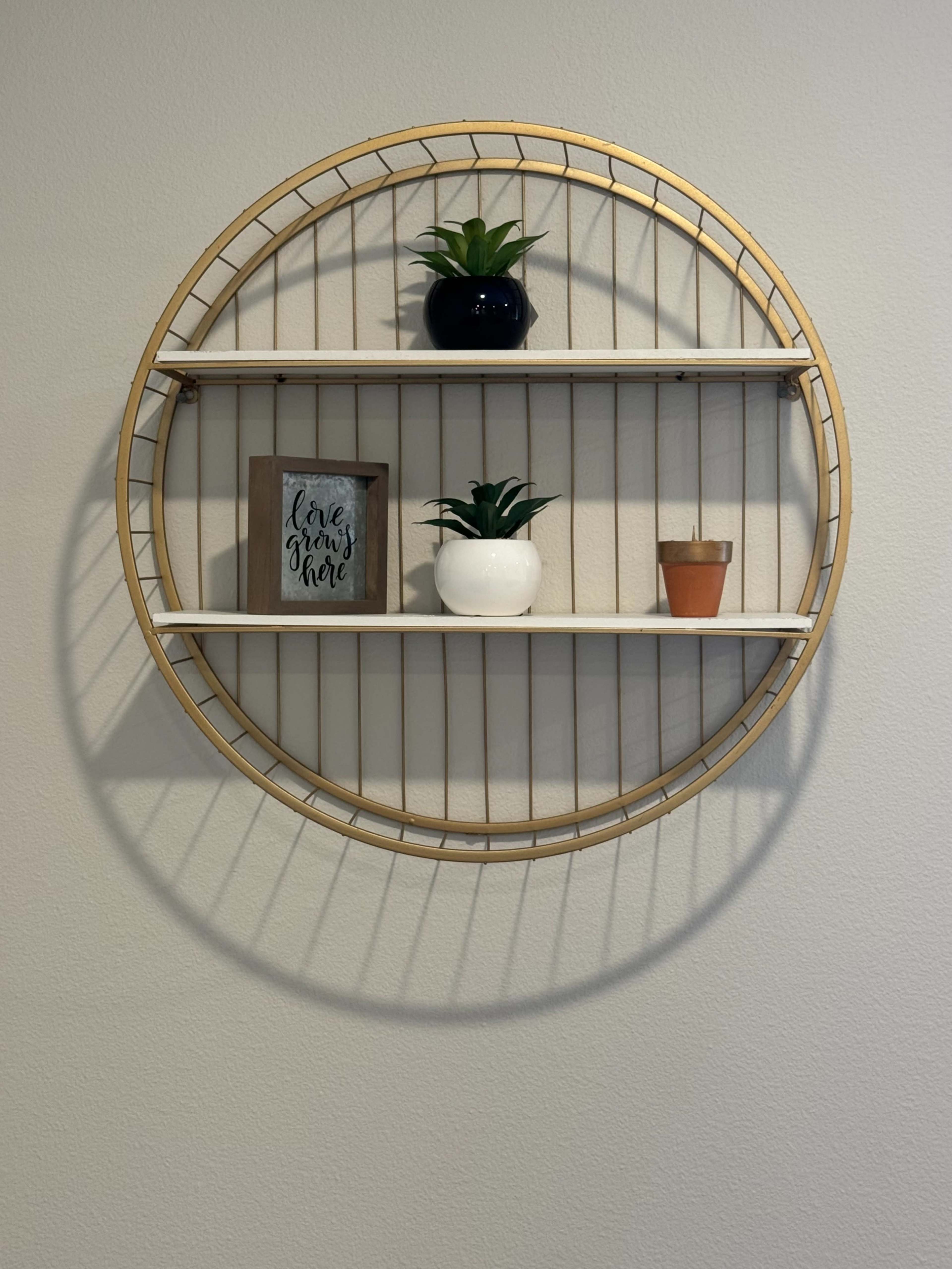 A circular wall shelf with two white shelves displays a framed sign, a small potted plant, and a decorative plant in a black pot against a light-colored wall.