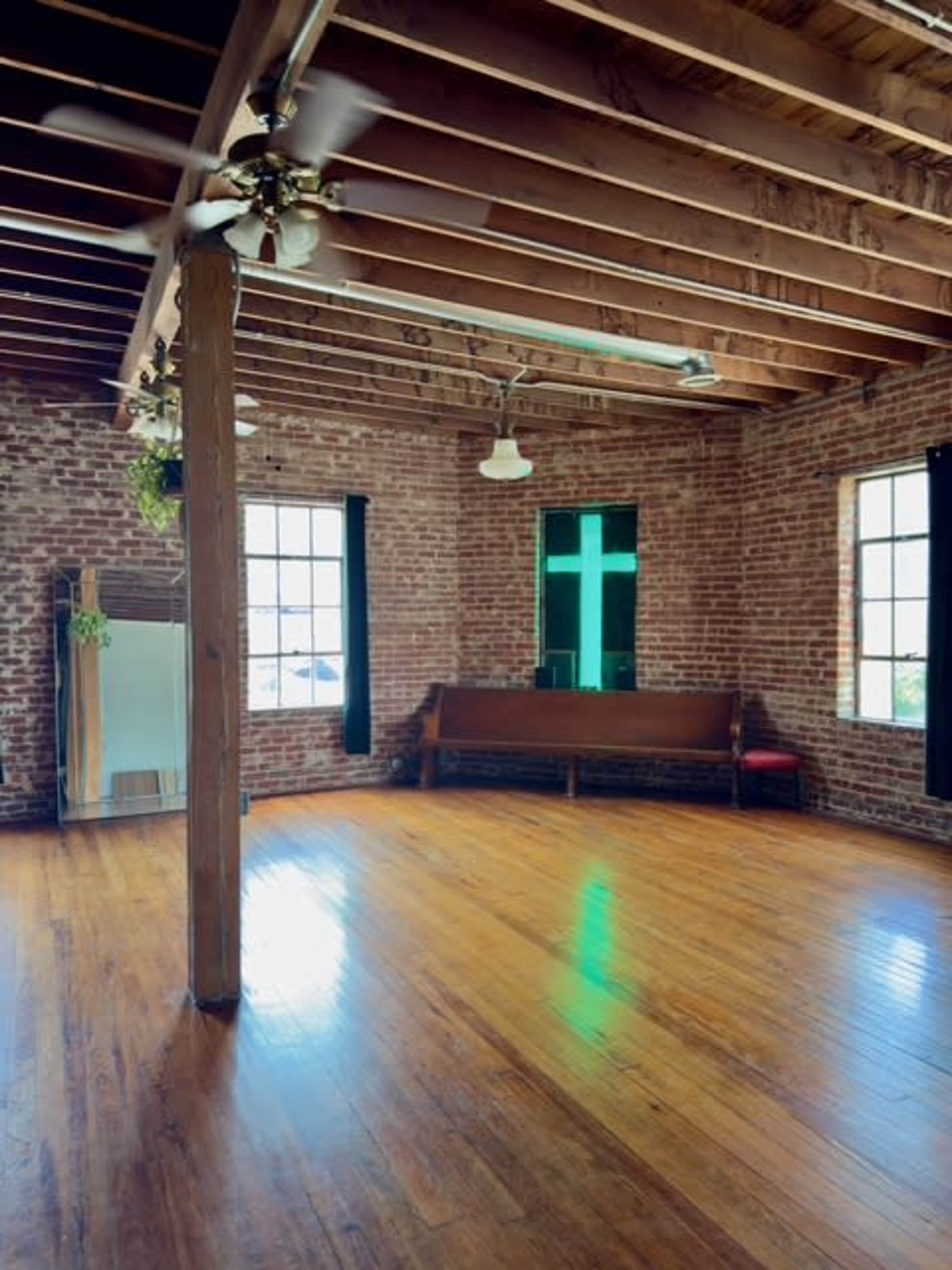The image shows an empty room with exposed wooden beams, brick walls, large windows, and polished wooden floors.