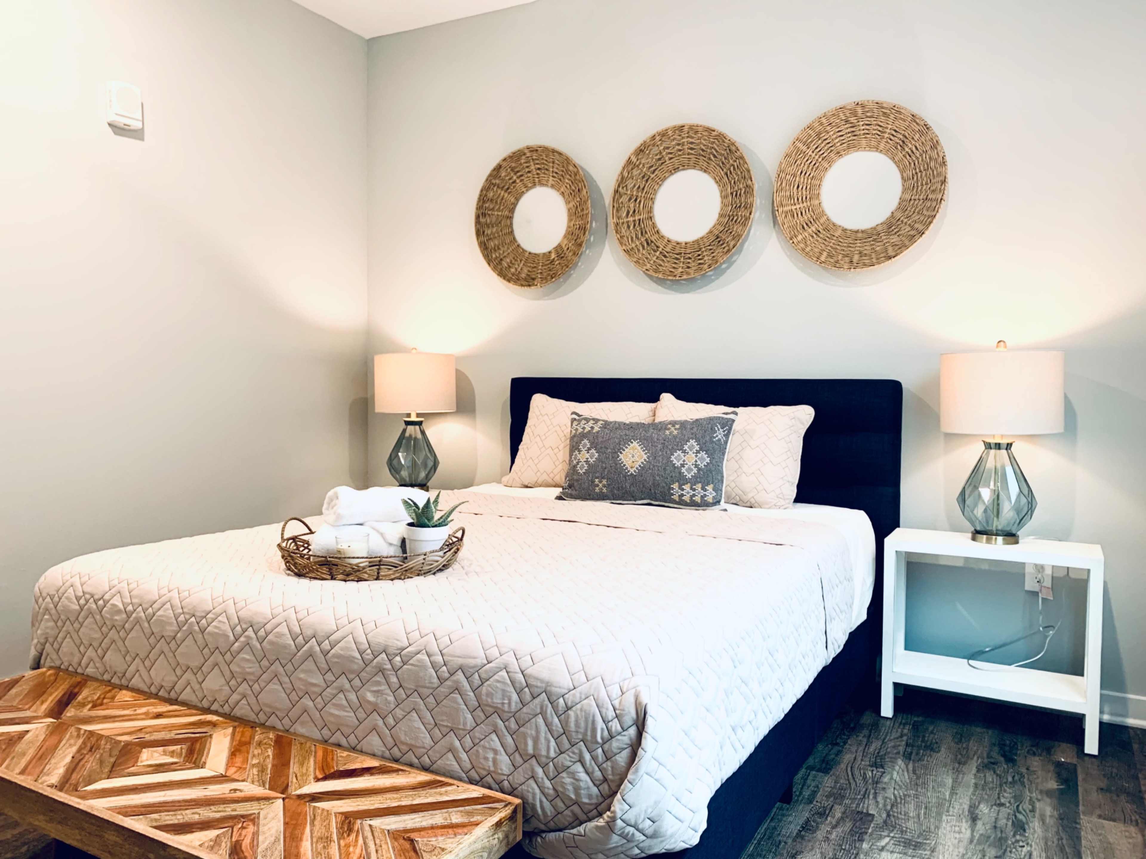 A neatly arranged bedroom features a bed with decorative pillows, two bedside lamps, wall art made of woven circles, and a wooden bench at the foot of the bed.