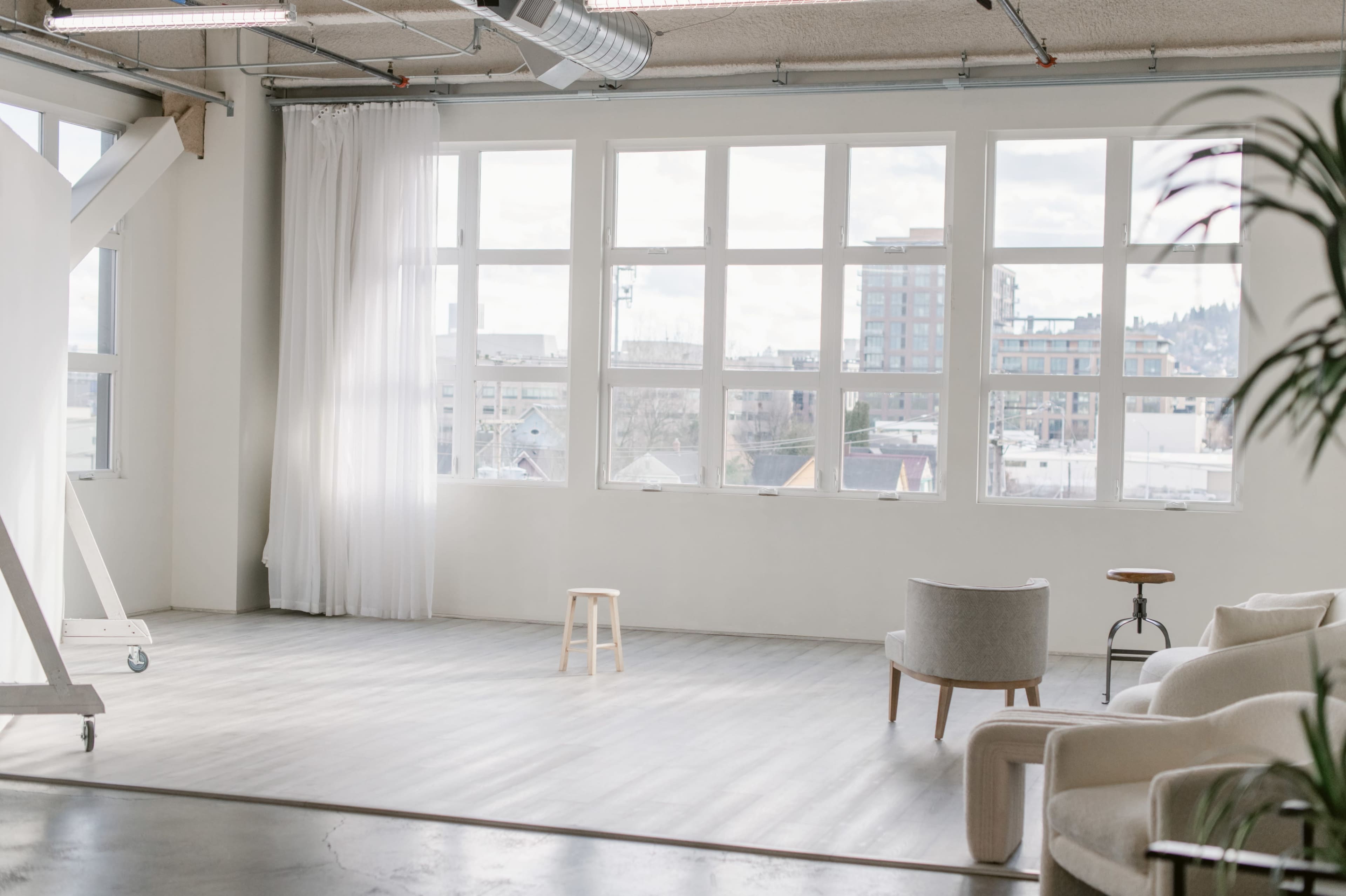 A spacious, well-lit room with large windows, a stool in the center, and a chair against the wall.