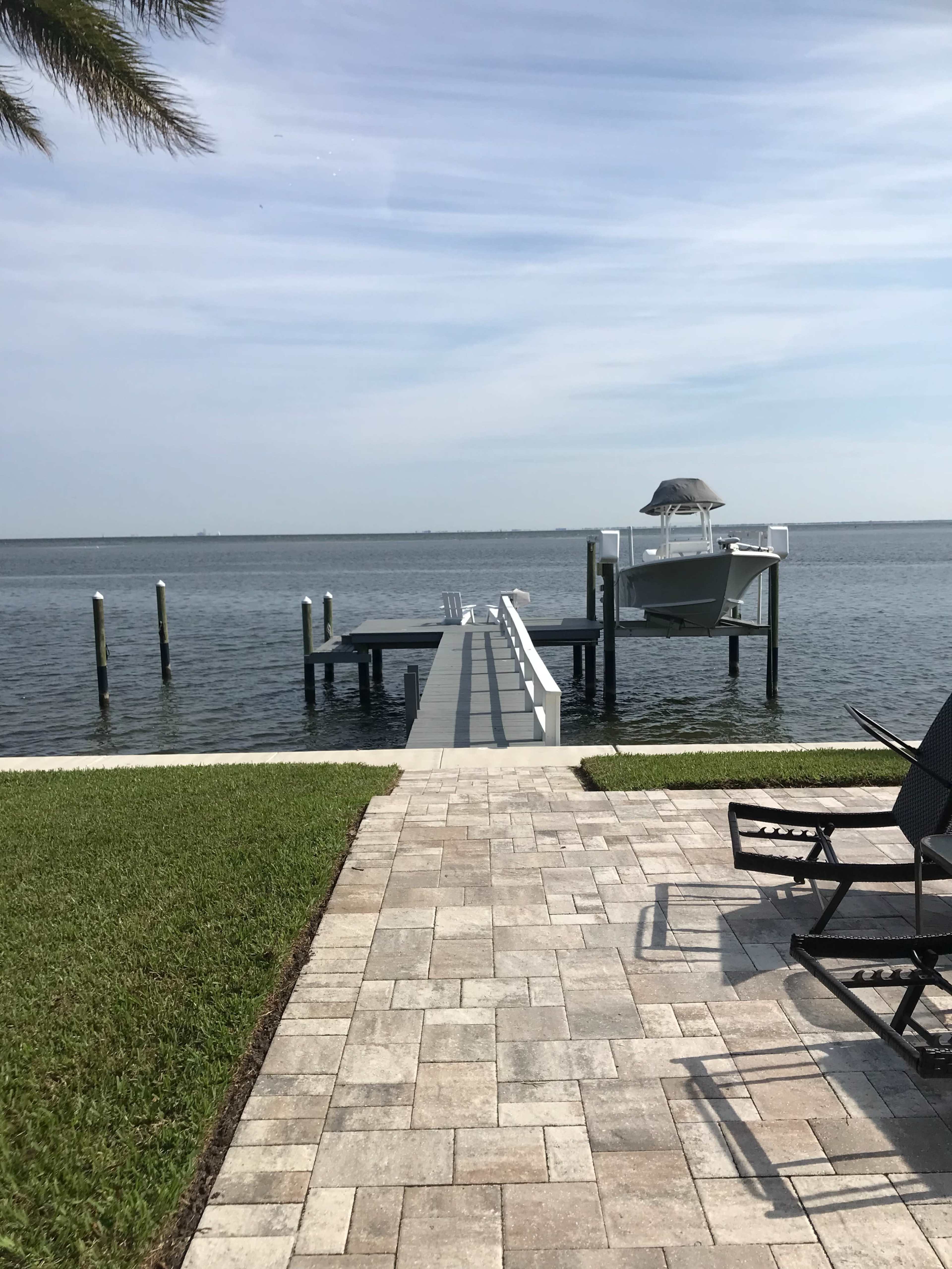 A wooden dock extends into calm water, with a boat moored at the end, framed by a neatly paved patio and a grassy area.