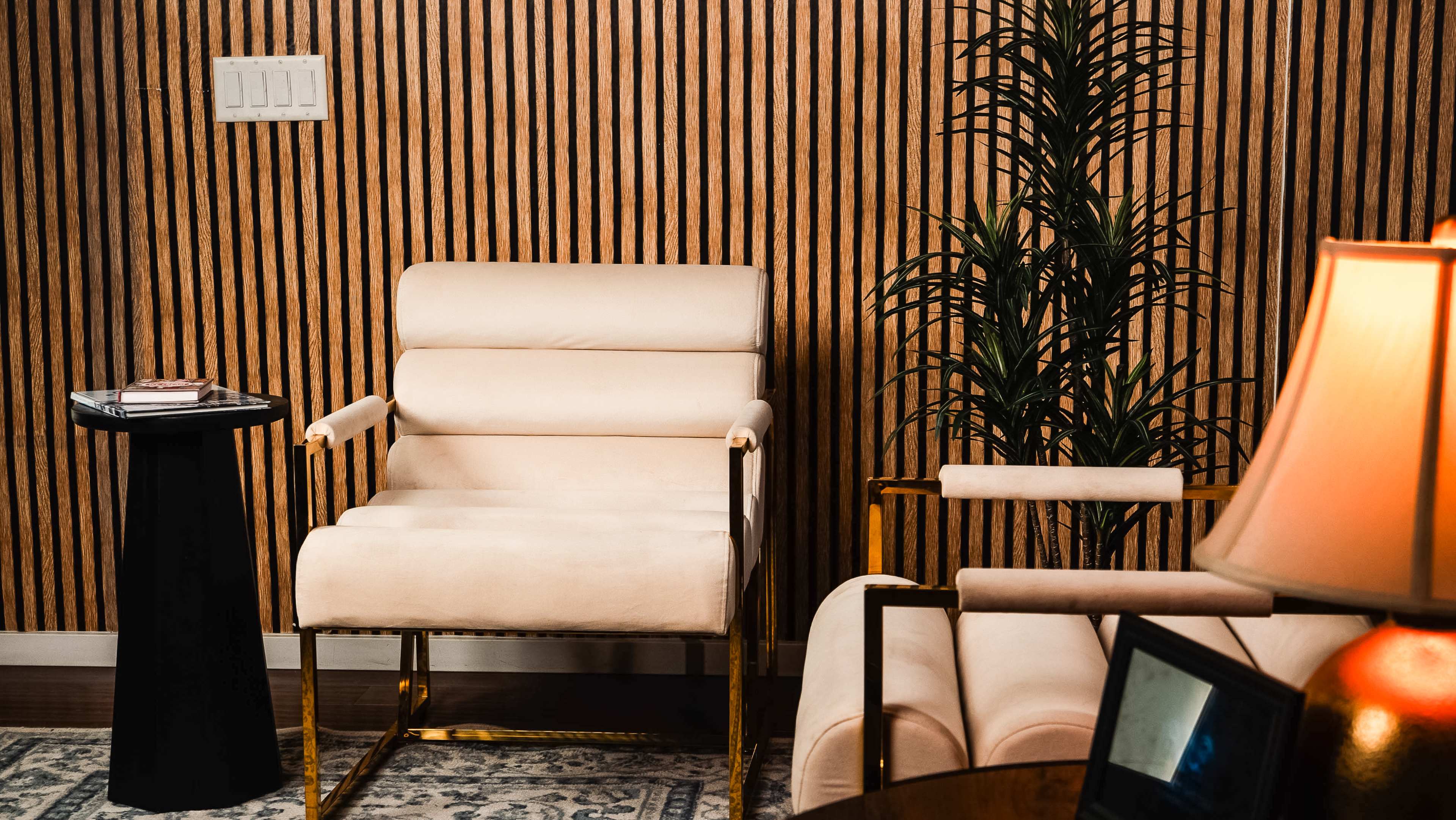 The image shows a minimalist seating area with two cream-colored chairs, a black side table, and a potted plant against a wall with vertical wood paneling.