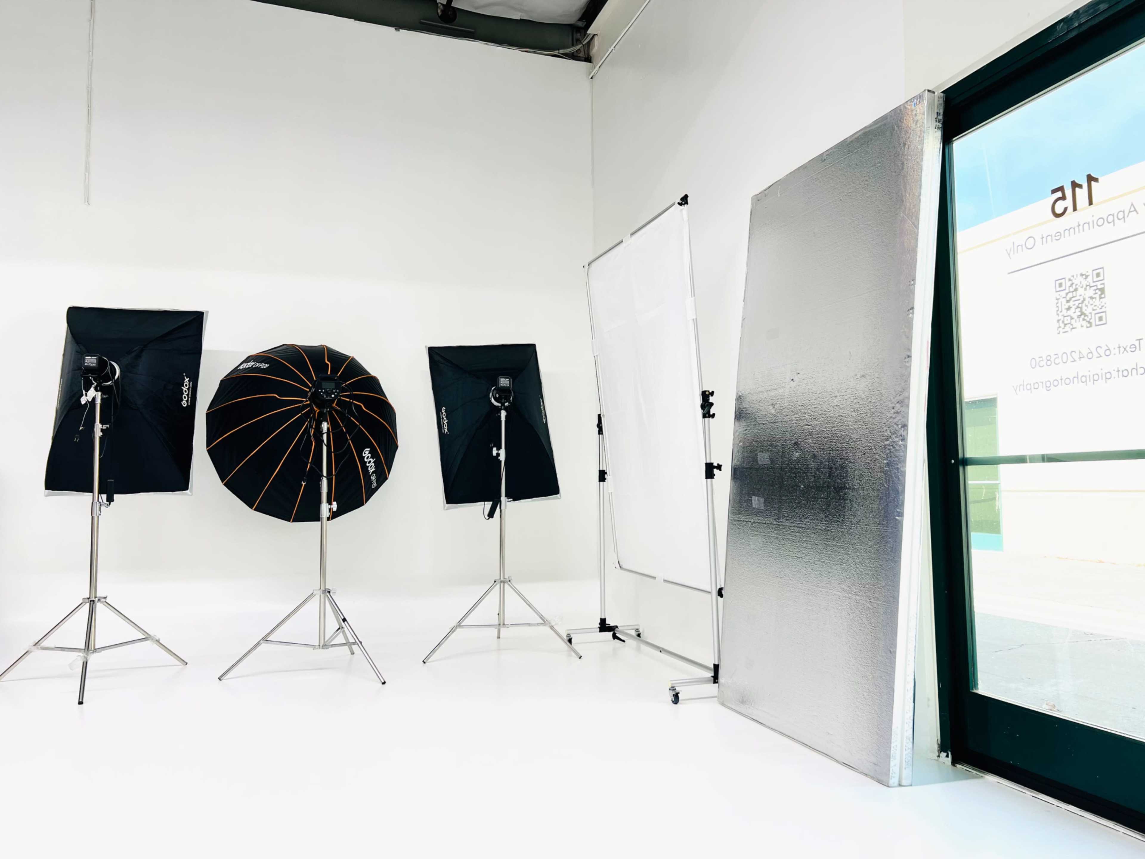 The image shows a bright studio space with three softbox light setups, a backdrop support frame, and a reflective panel.