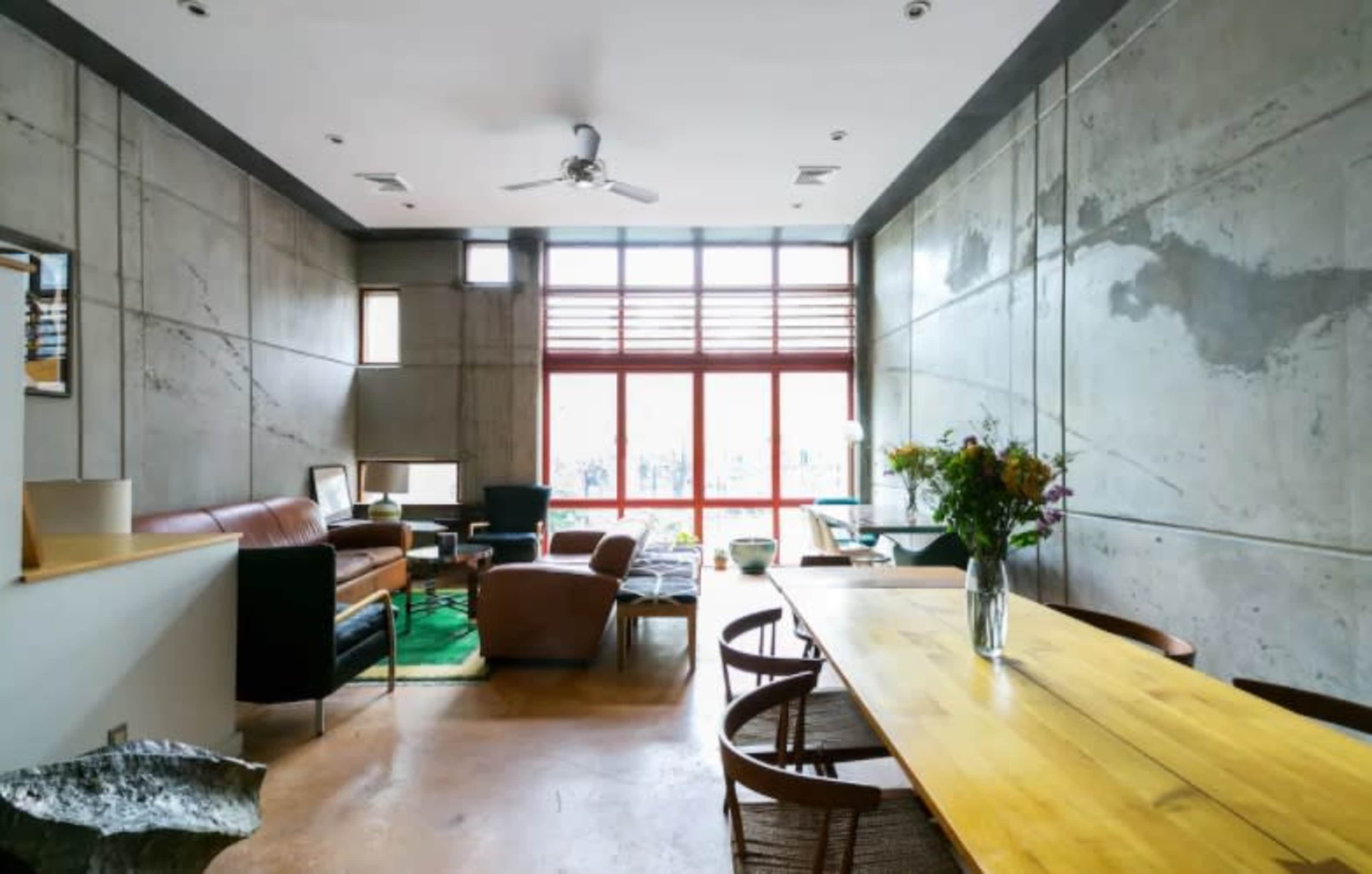 A modern living space with concrete walls, a wooden dining table, and large windows allowing natural light to enter.