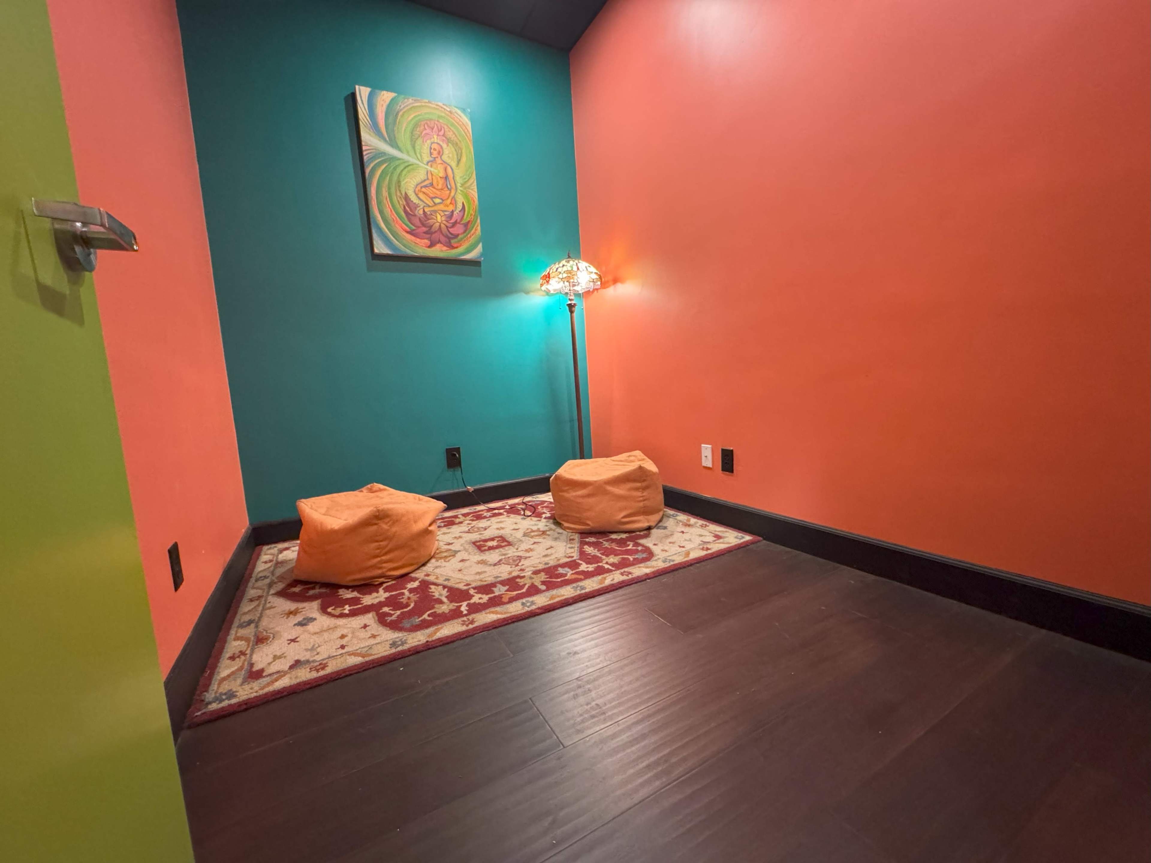 The image shows a small, brightly colored room with an orange and teal wall, featuring a rug, two beanbag chairs, and a lamp.