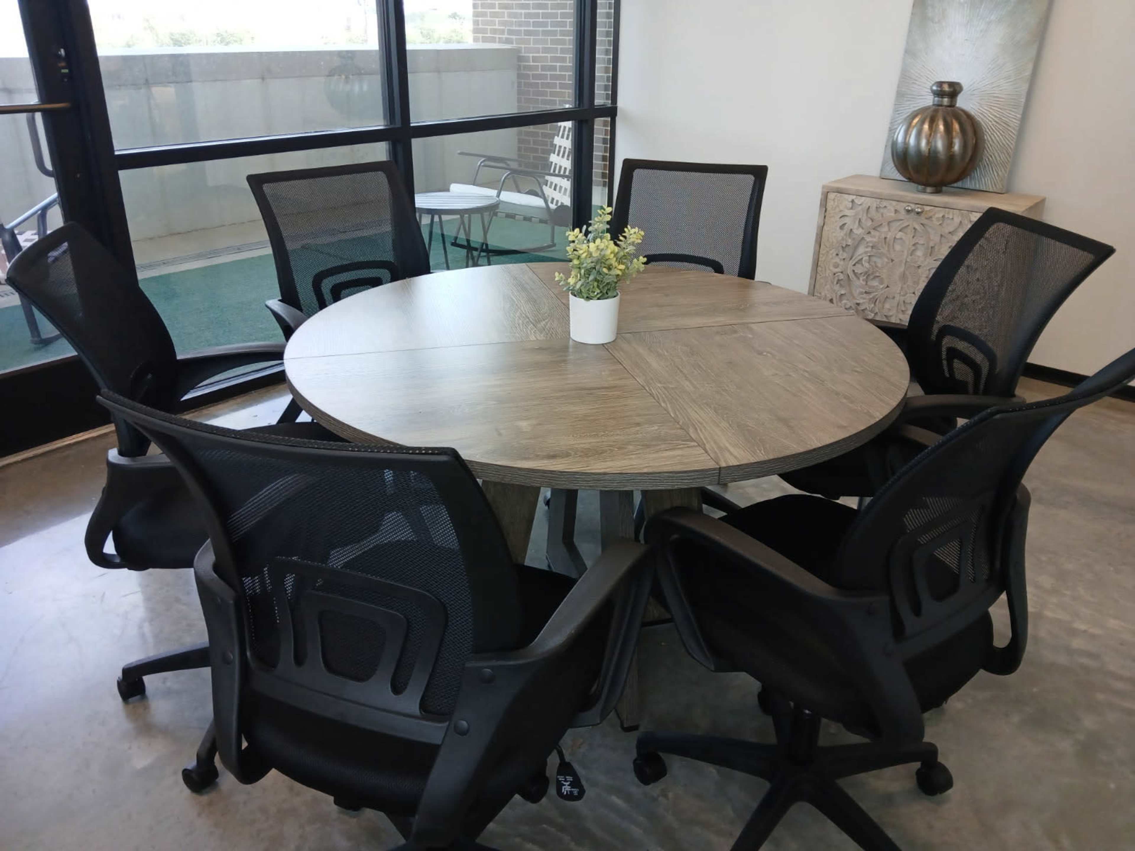 A round wooden conference table surrounded by six black mesh office chairs is positioned near large windows.