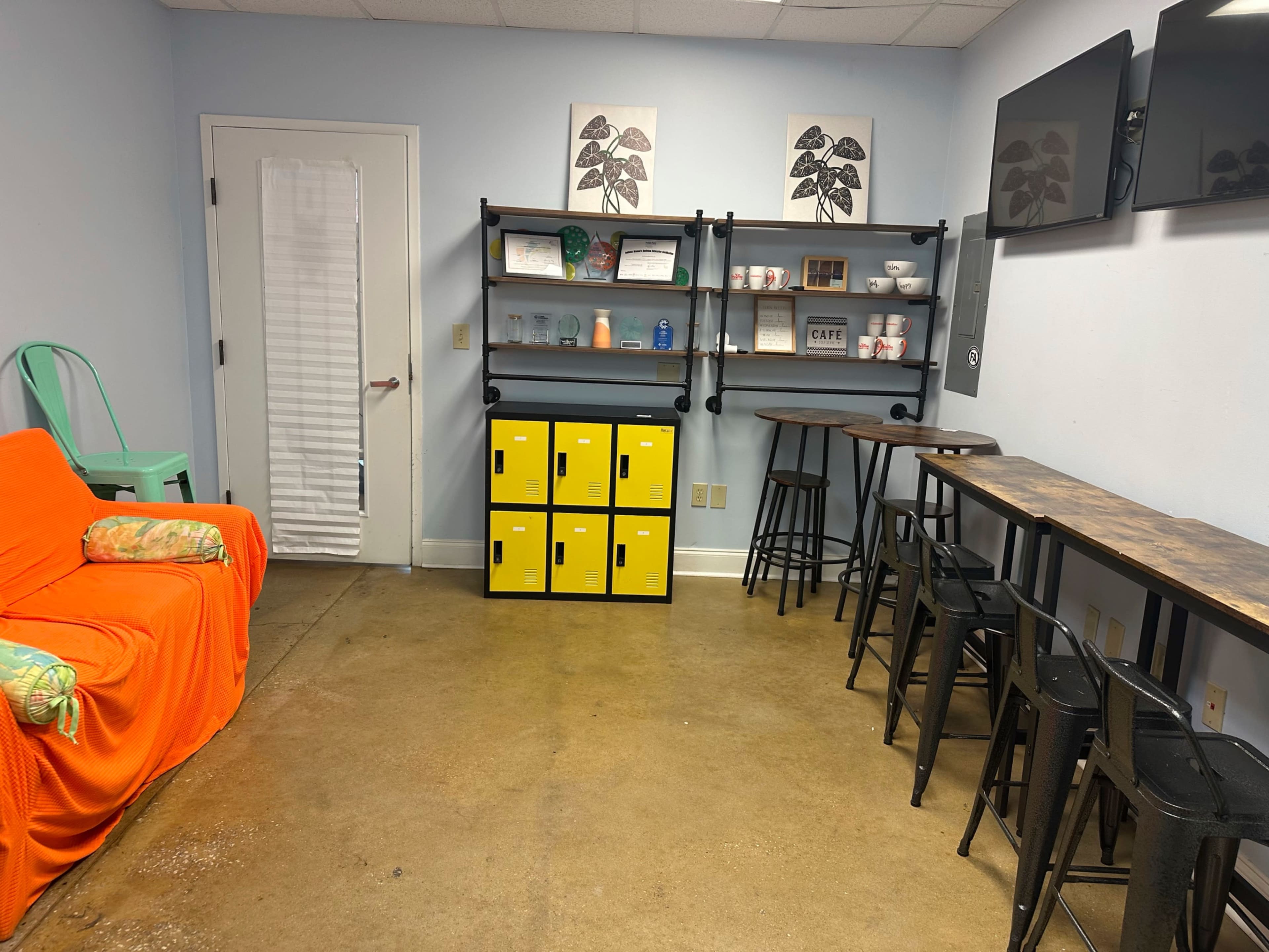 The image shows a small break room with a bright orange couch, shelving unit with various items, yellow storage bins, and high-top tables with black stools.