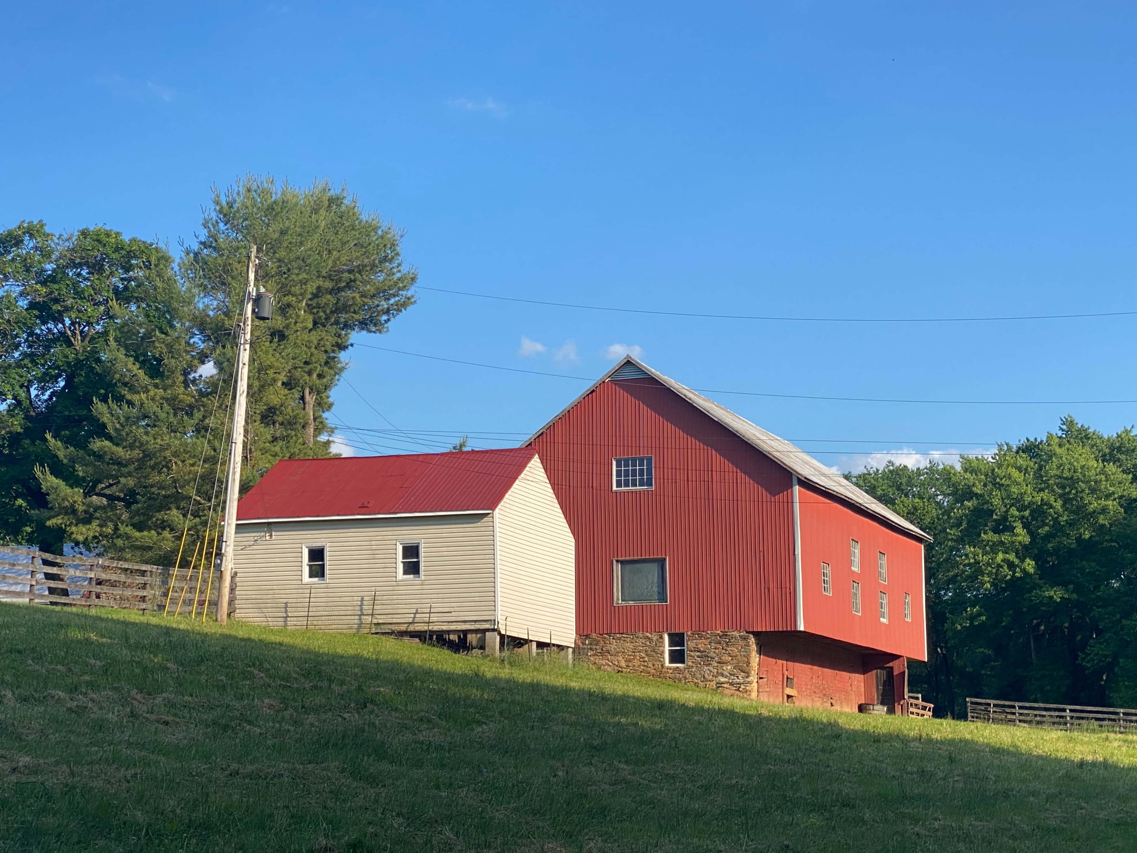 A red barn and a light-colored building with a sloped roof situated on a grassy hillside surrounded by trees.