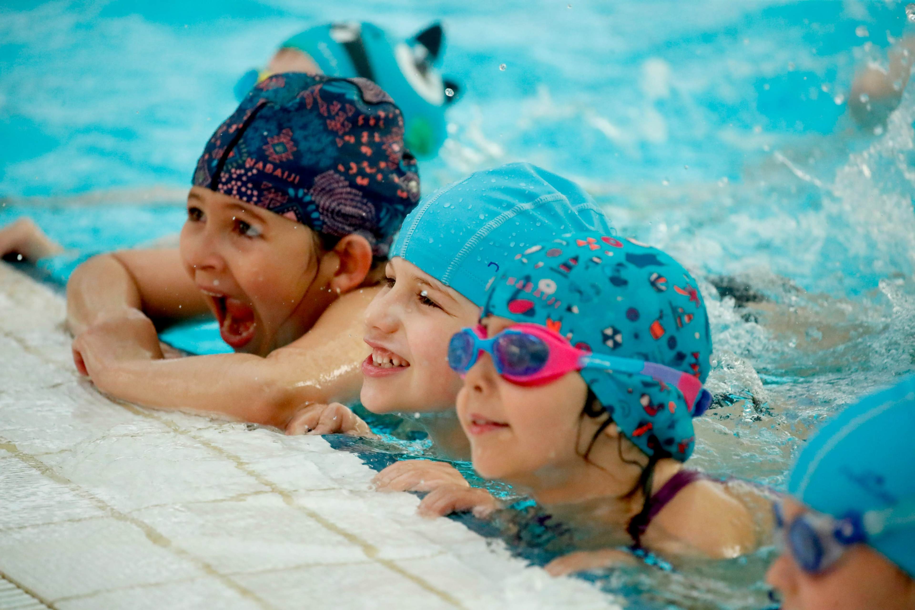 A group of children in swim caps and goggles are smiling and leaning against the edge of a pool.