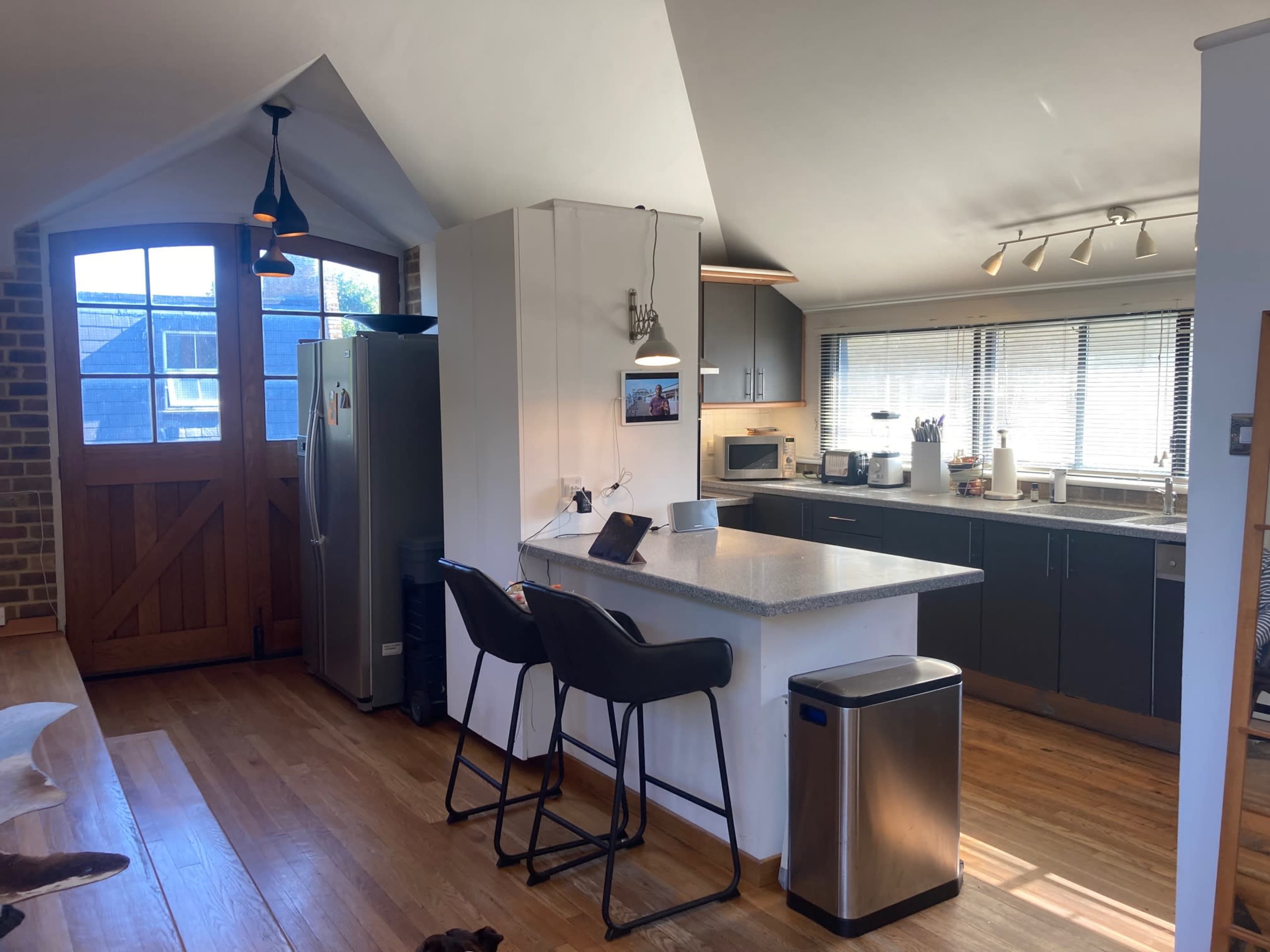 The image shows a modern kitchen with a central island, equipped with bar stools, stainless steel appliances, and large windows that provide natural light.