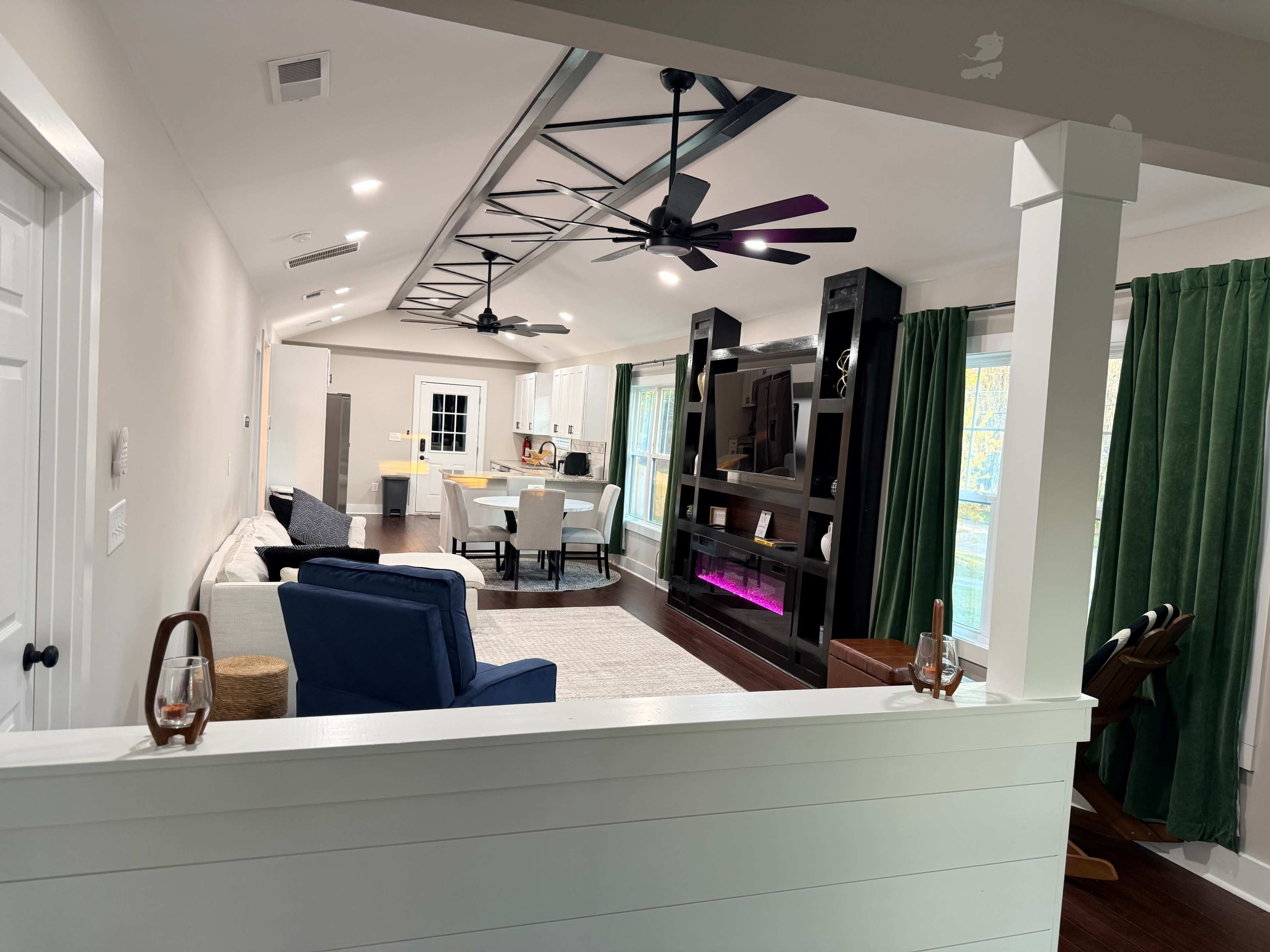 The image shows a modern living space with a sofa, dining area, and a black entertainment center, lit by natural light through large windows and accented by green curtains.