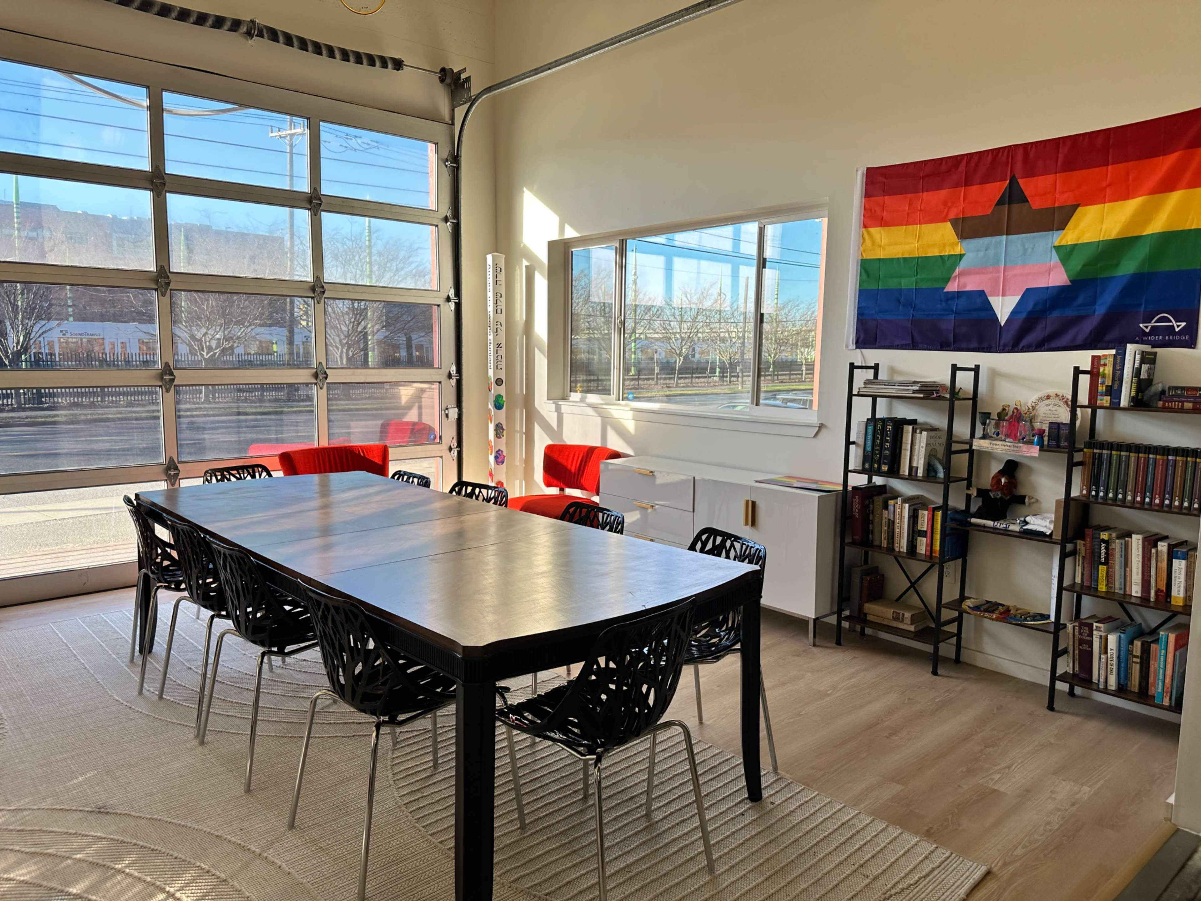 A bright room features a large black table surrounded by black chairs, with a colorful flag on the wall and a bookshelf filled with books in the corner.