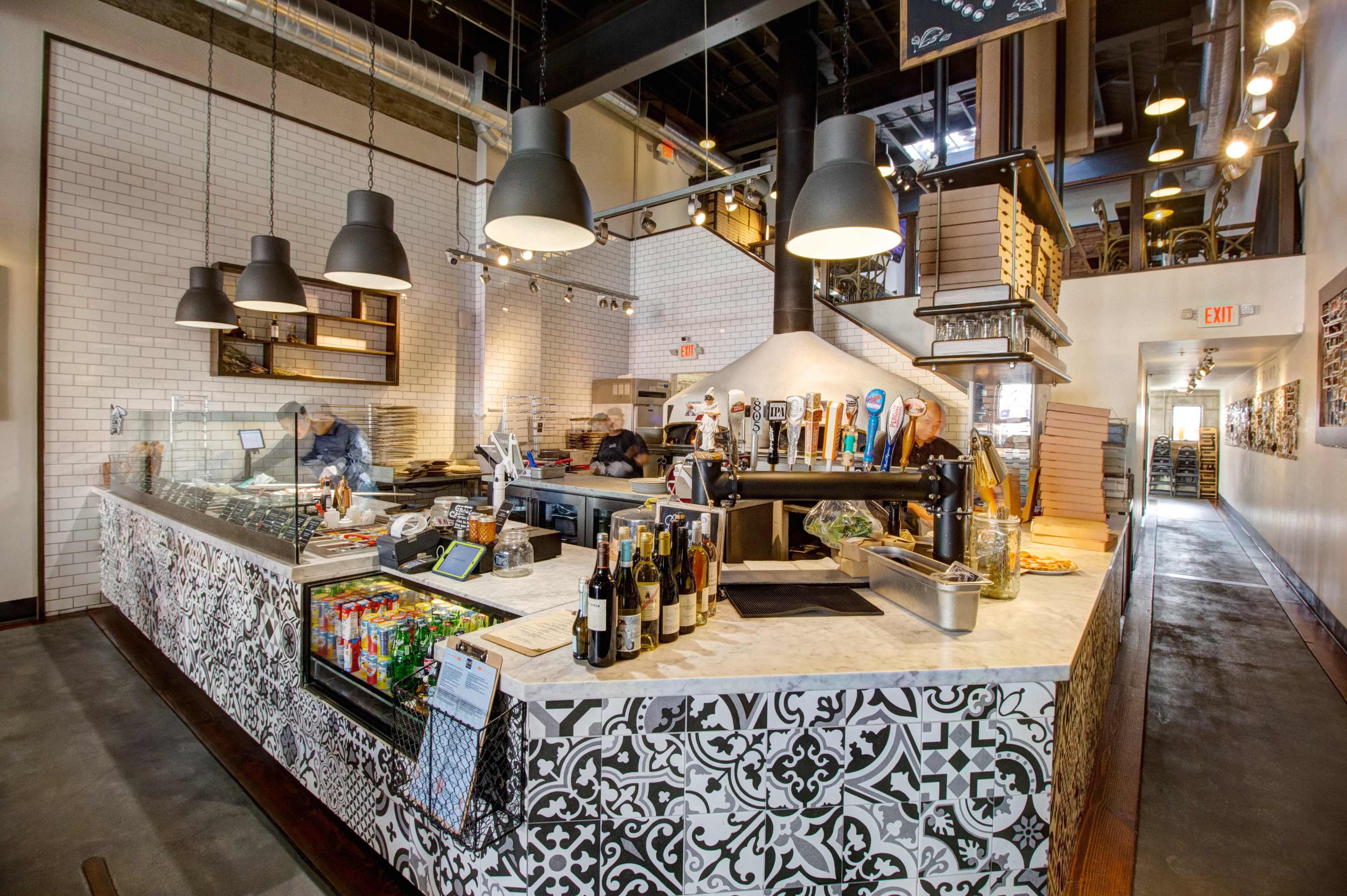 A modern restaurant interior with a bar area featuring various drinks, a kitchen space where staff are preparing food, and decorative tile work on the counter.