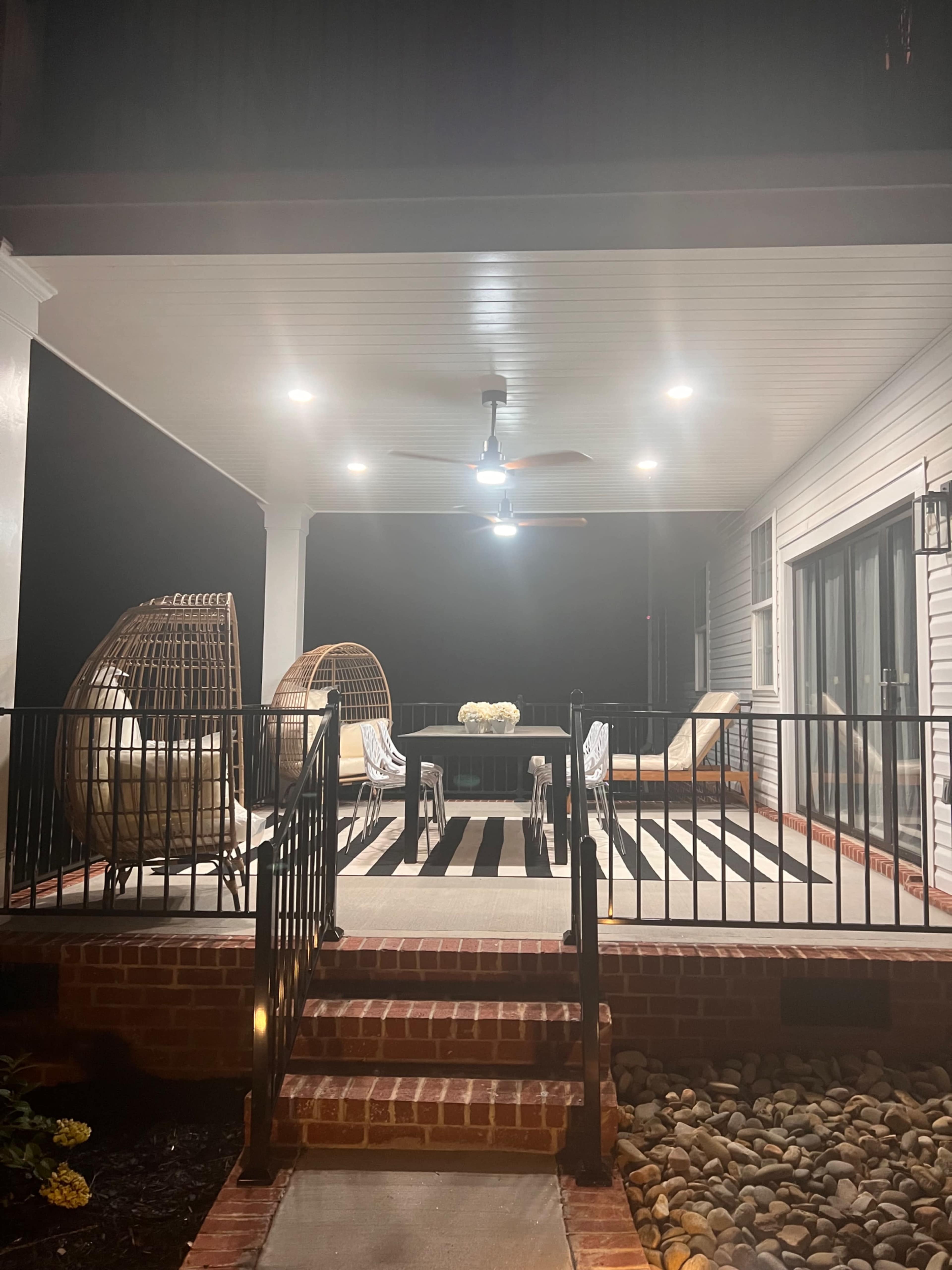 A covered outdoor patio with two wicker chairs, a table with a floral centerpiece, and a ceiling fan, illuminated by overhead lights on a dark night.