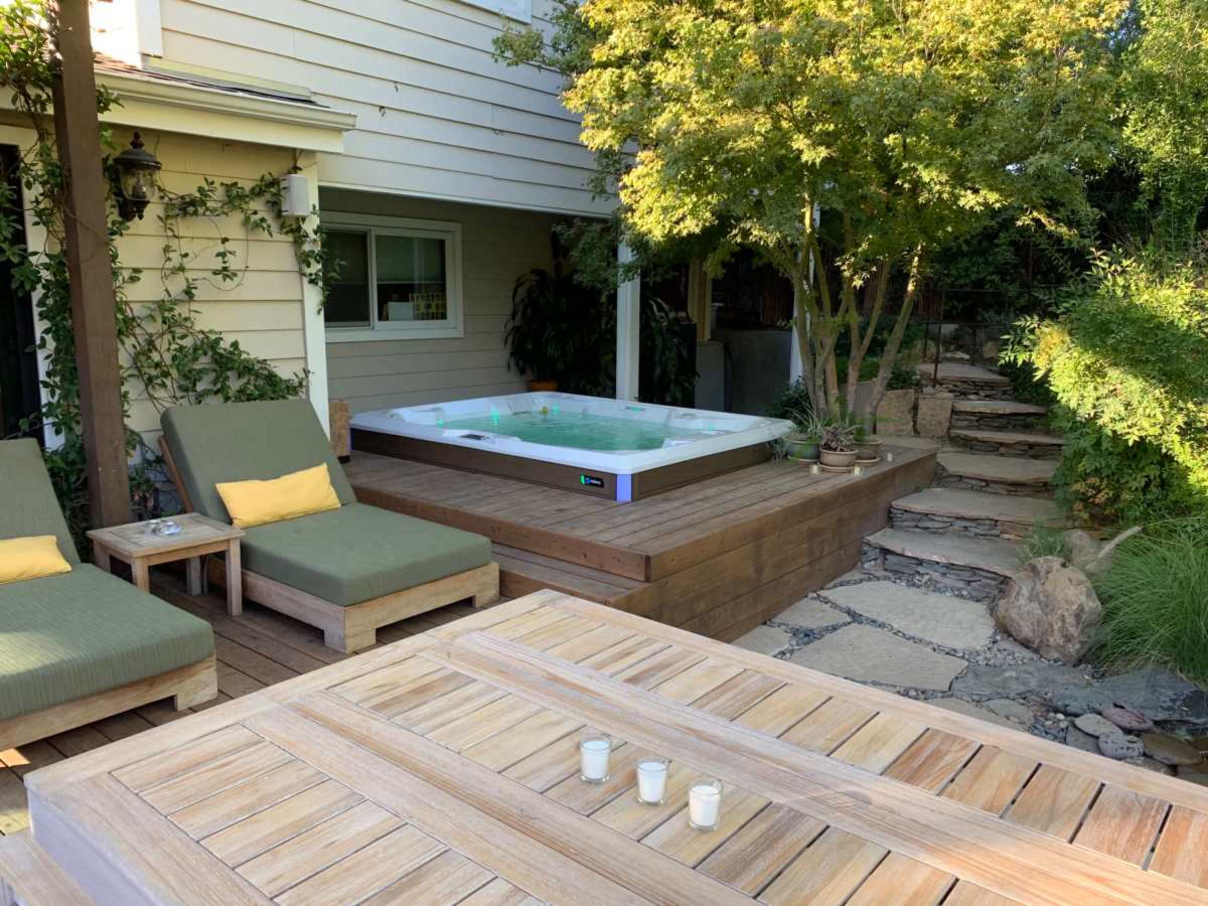 A hot tub is situated on a wooden deck surrounded by greenery, lounge chairs, and a stone pathway leading to a garden area.