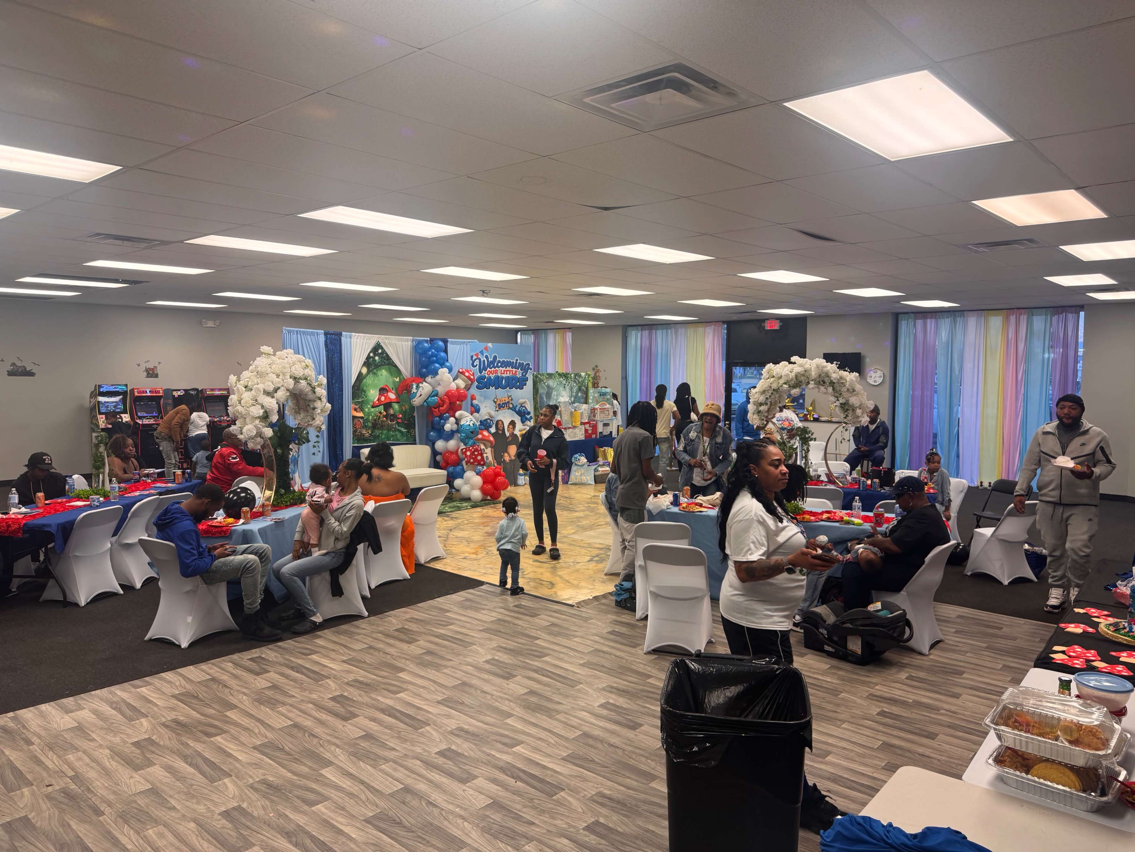 The image shows a lively indoor event space decorated with balloons and table settings, where people are engaged in conversations and activities.