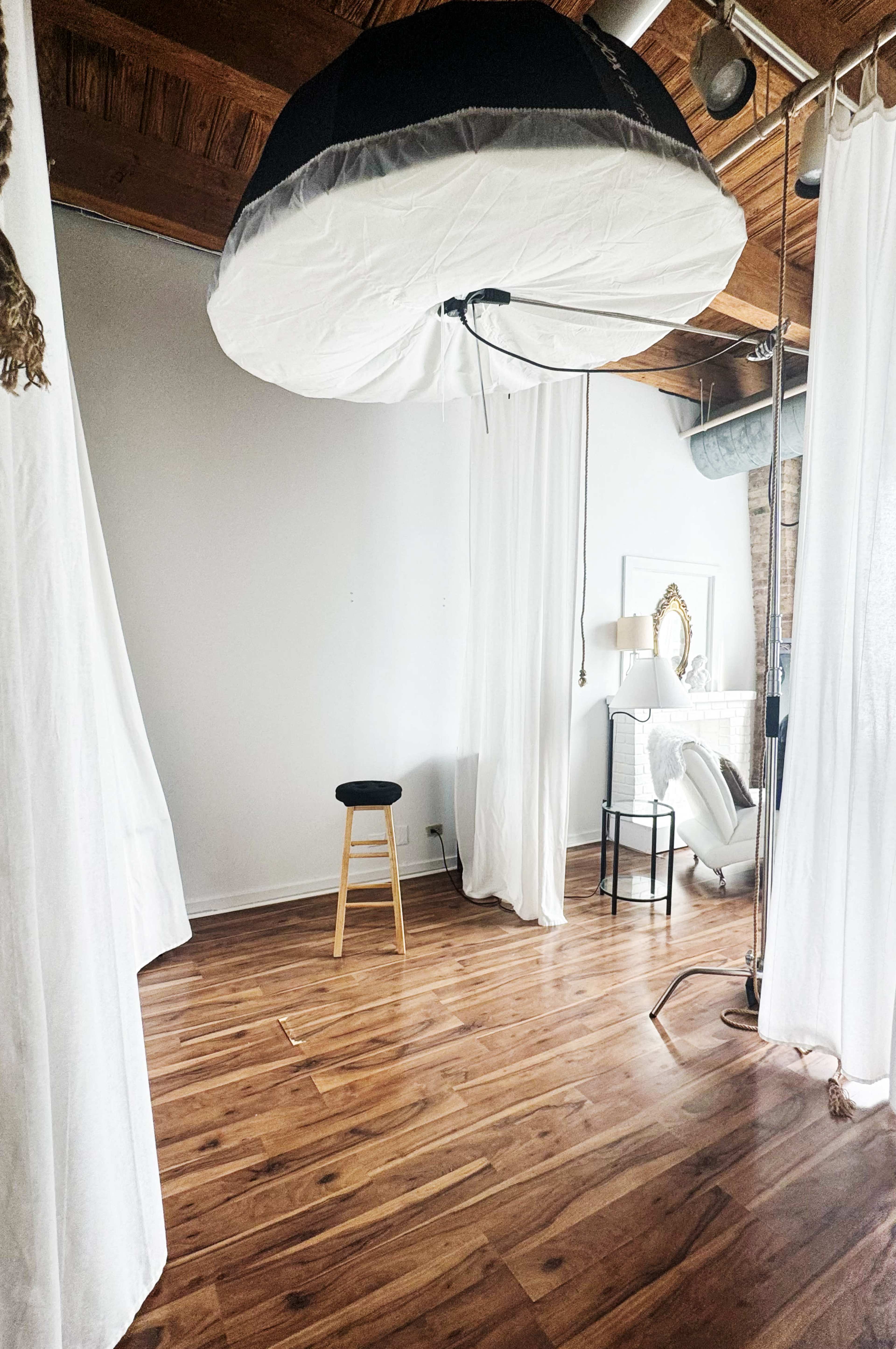 The image shows a photography studio with a wooden floor, white curtains, a stool, and a large softbox light hanging from the ceiling.