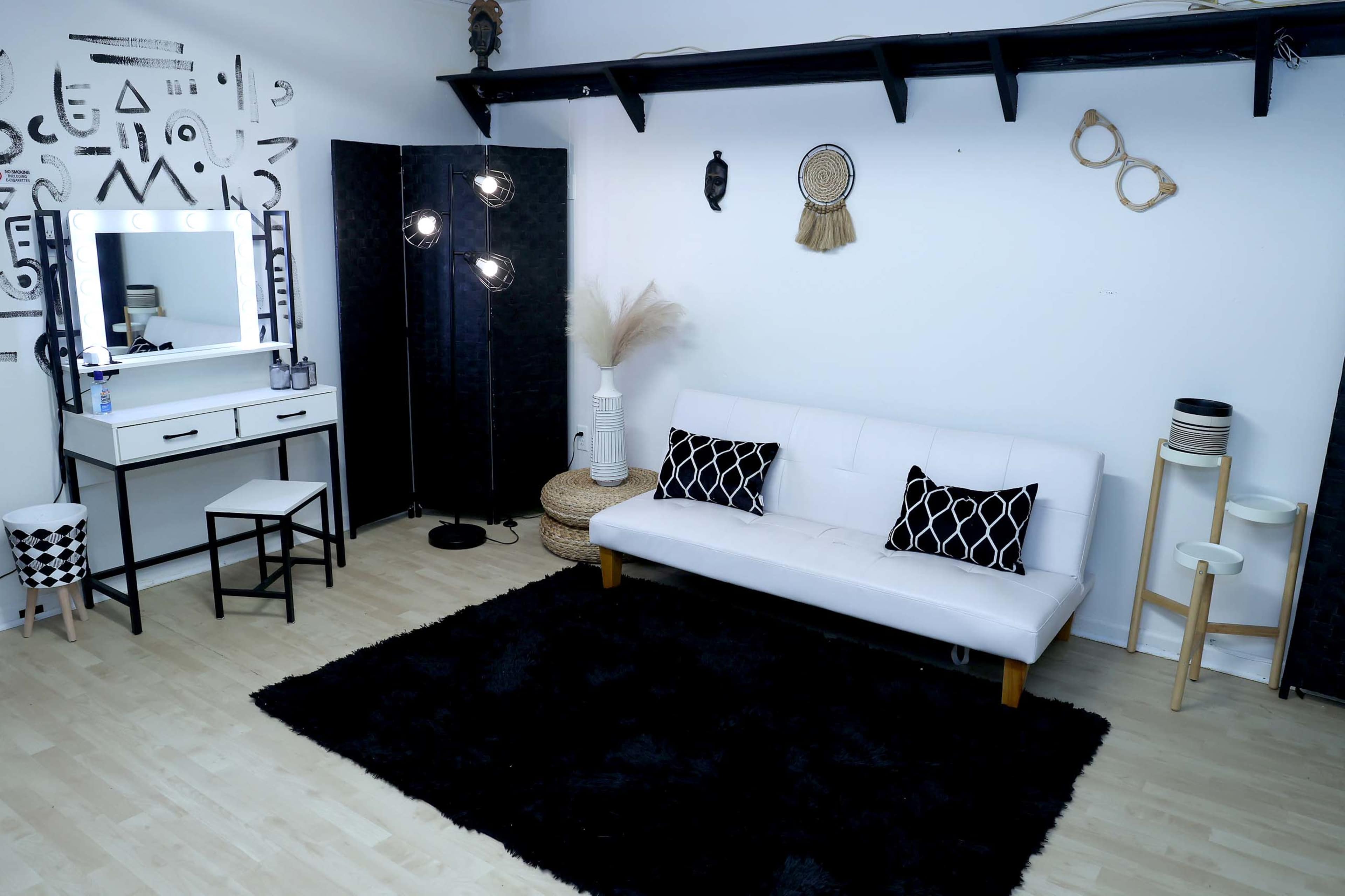 The room features a white sofa with two black and white decorative pillows, a black area rug, a modern vanity with a mirror, and various decorative items against a plain white wall.
