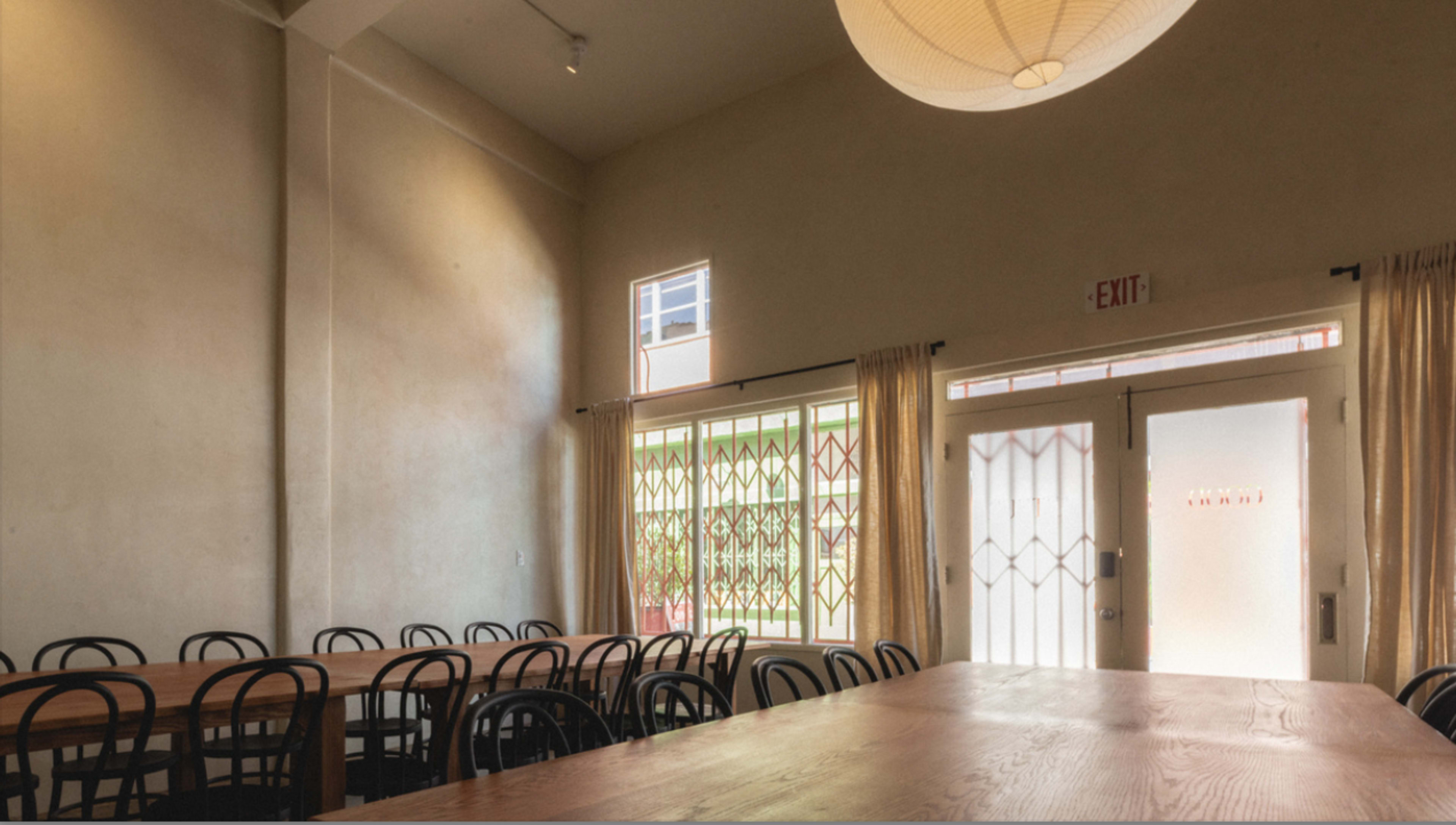 The image shows a dining area with a long wooden table and black chairs, featuring large windows and a door at one end.