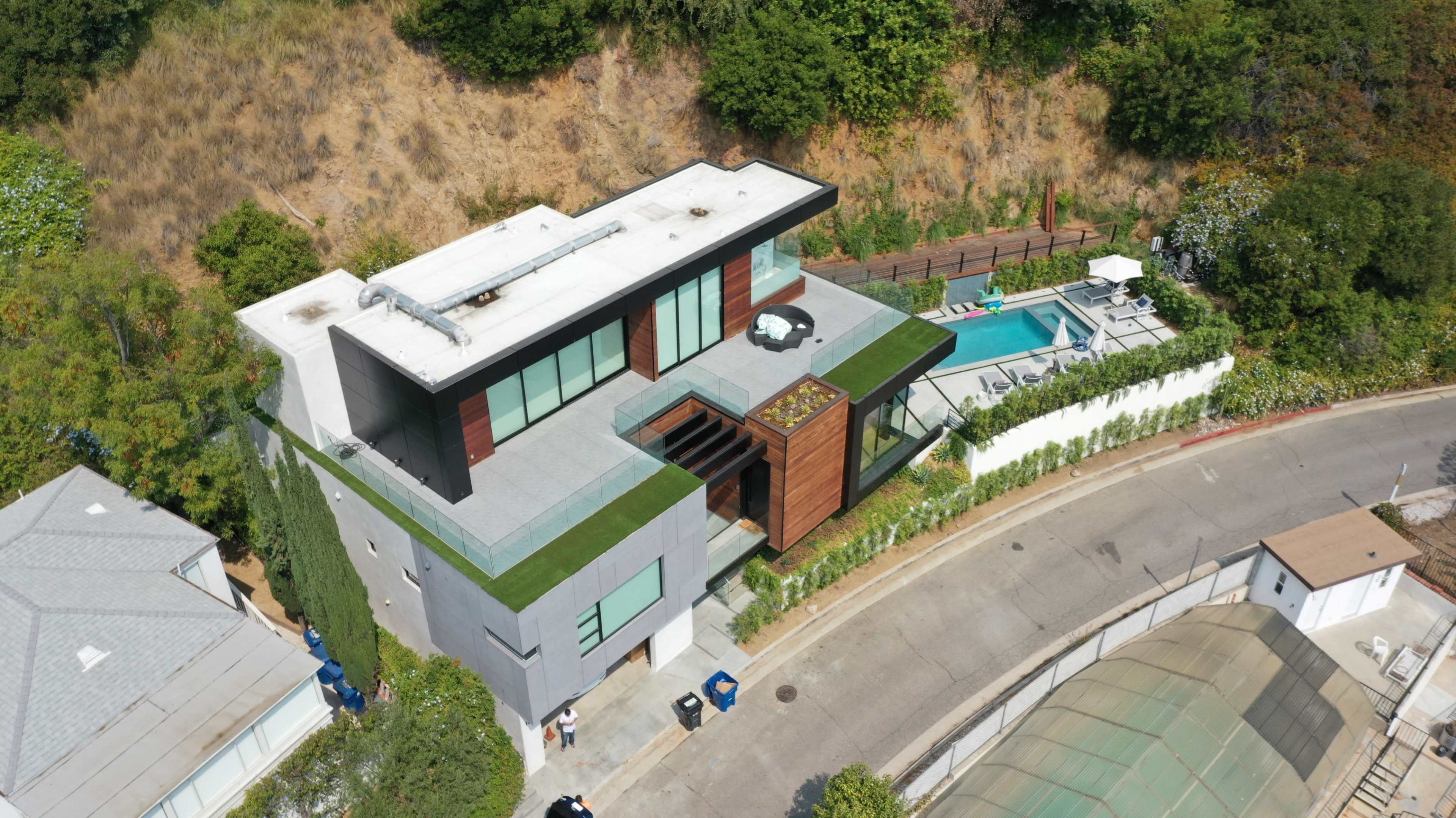 A modern house with a flat roof and green landscaping, situated on a hillside near a swimming pool and a winding road.
