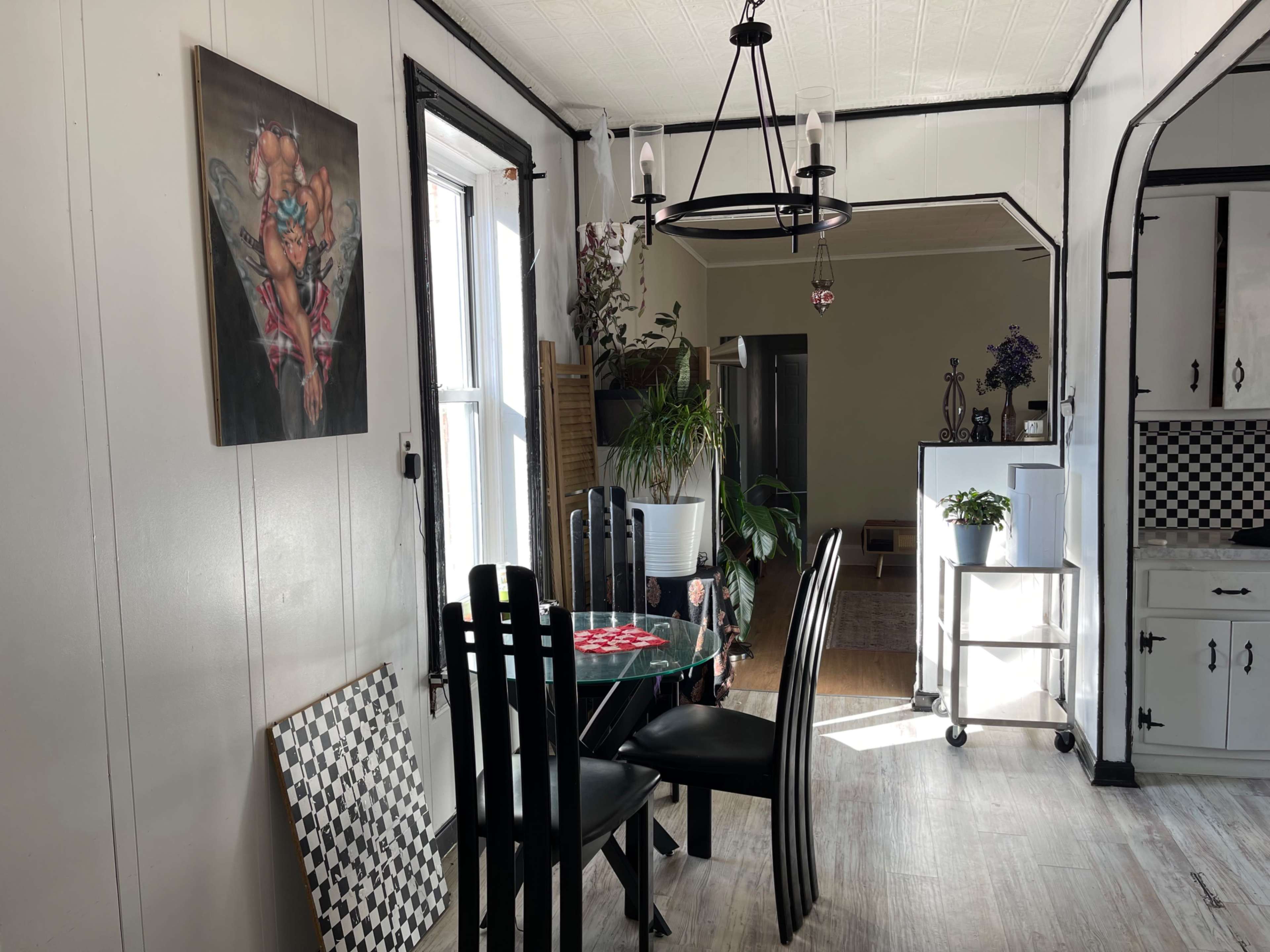 The image shows a dining area with a round glass table surrounded by black chairs, a piece of artwork on the wall, and a large window allowing natural light to enter.
