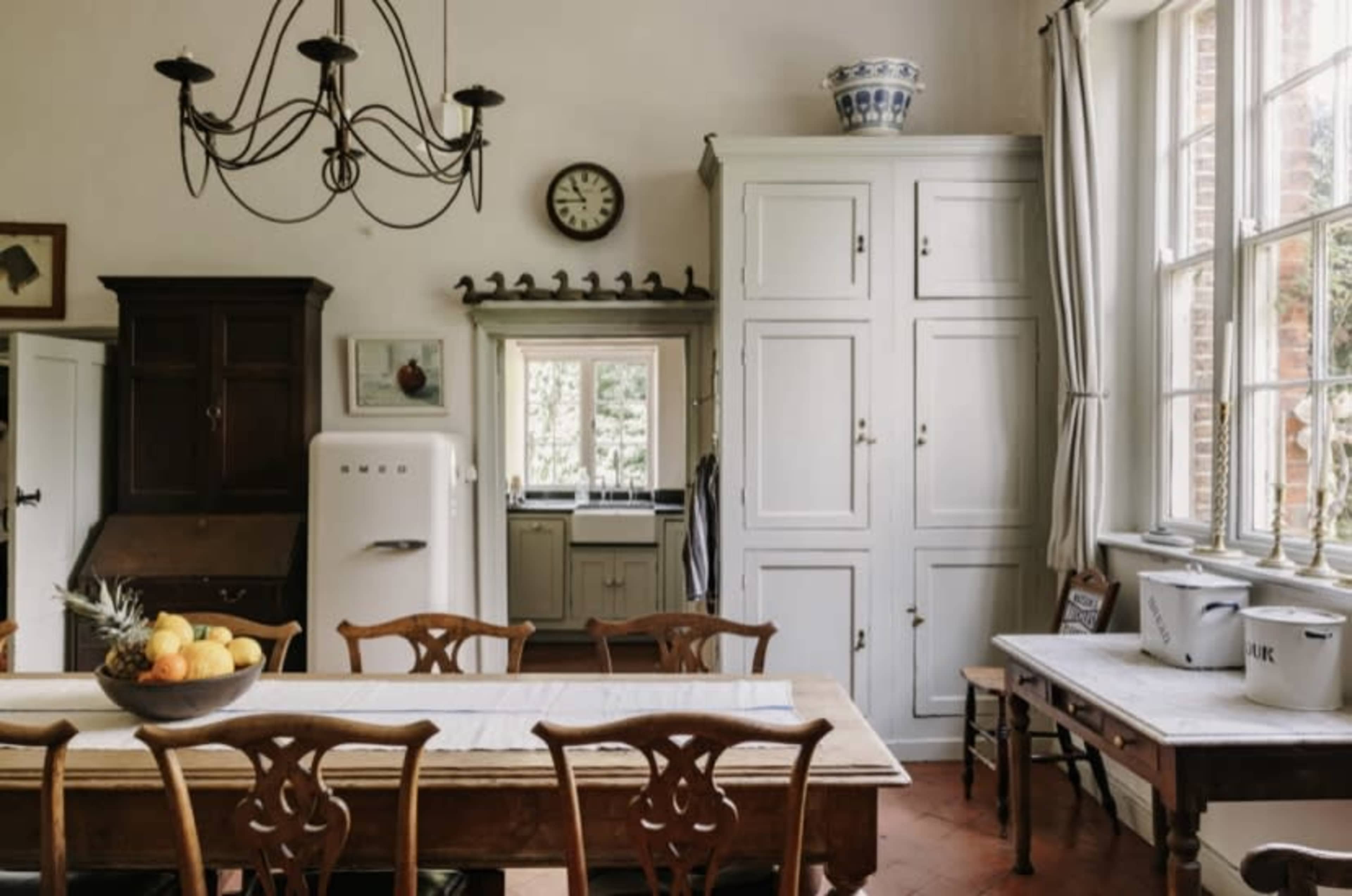 A well-lit kitchen features a large wooden table, a vintage refrigerator, and a white cabinet, with windows providing a view of the outdoors.