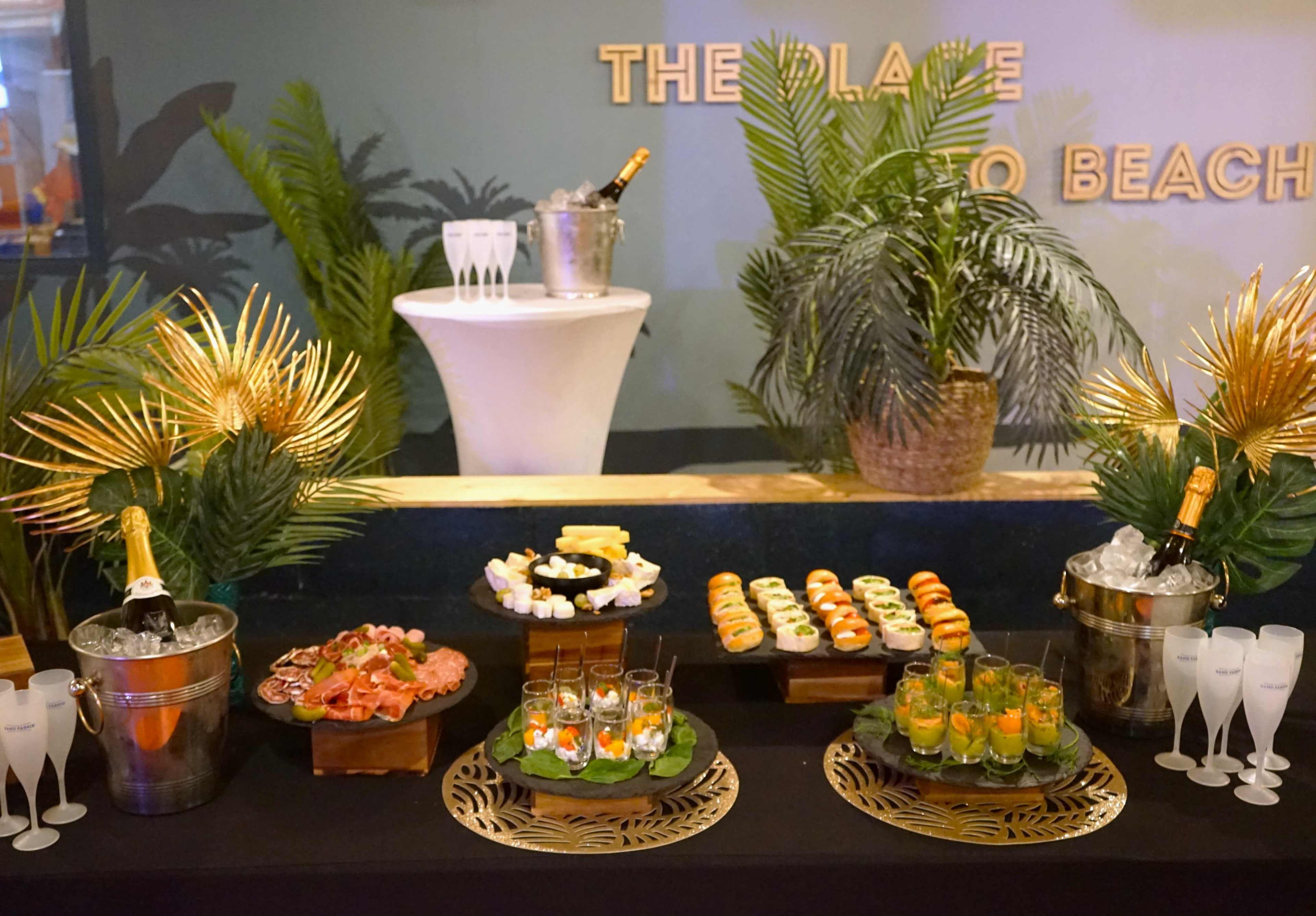 A buffet display features a variety of sushi, seafood, and cocktails set against a backdrop of decorative plants and a champagne bucket.