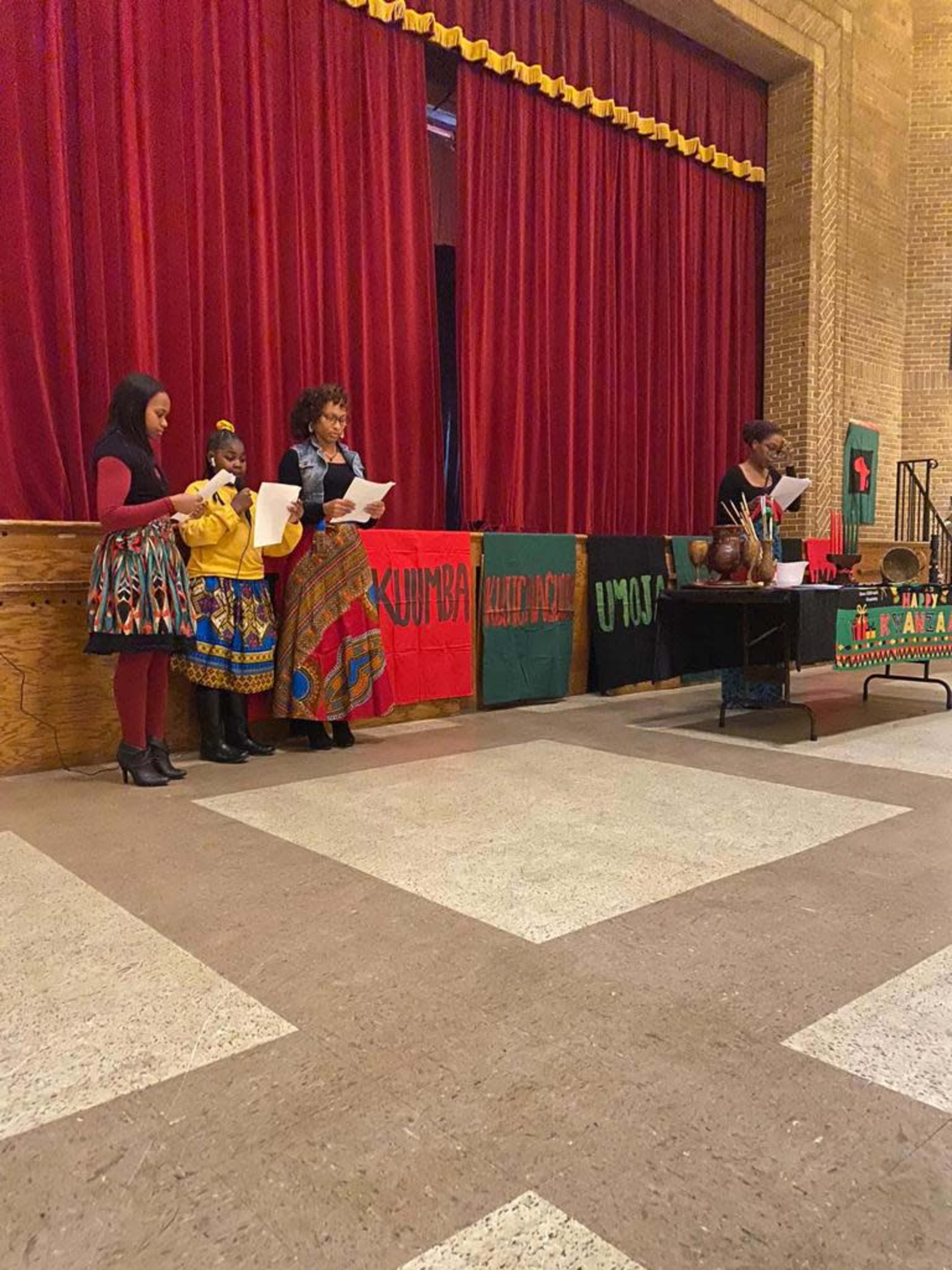 A group of individuals stands in front of a red curtain, engaged in a presentation, with cultural attire and banners visible in the background.