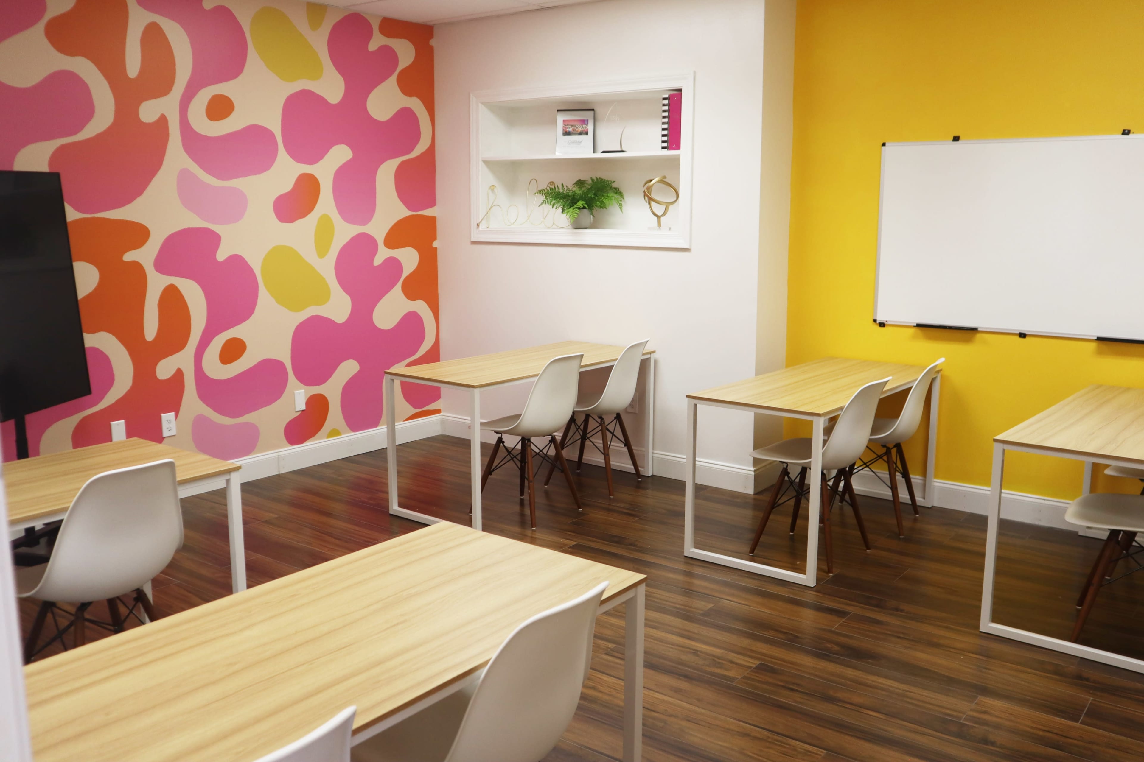 A brightly colored classroom with six wooden tables and chairs, set against a pink and yellow patterned wall.