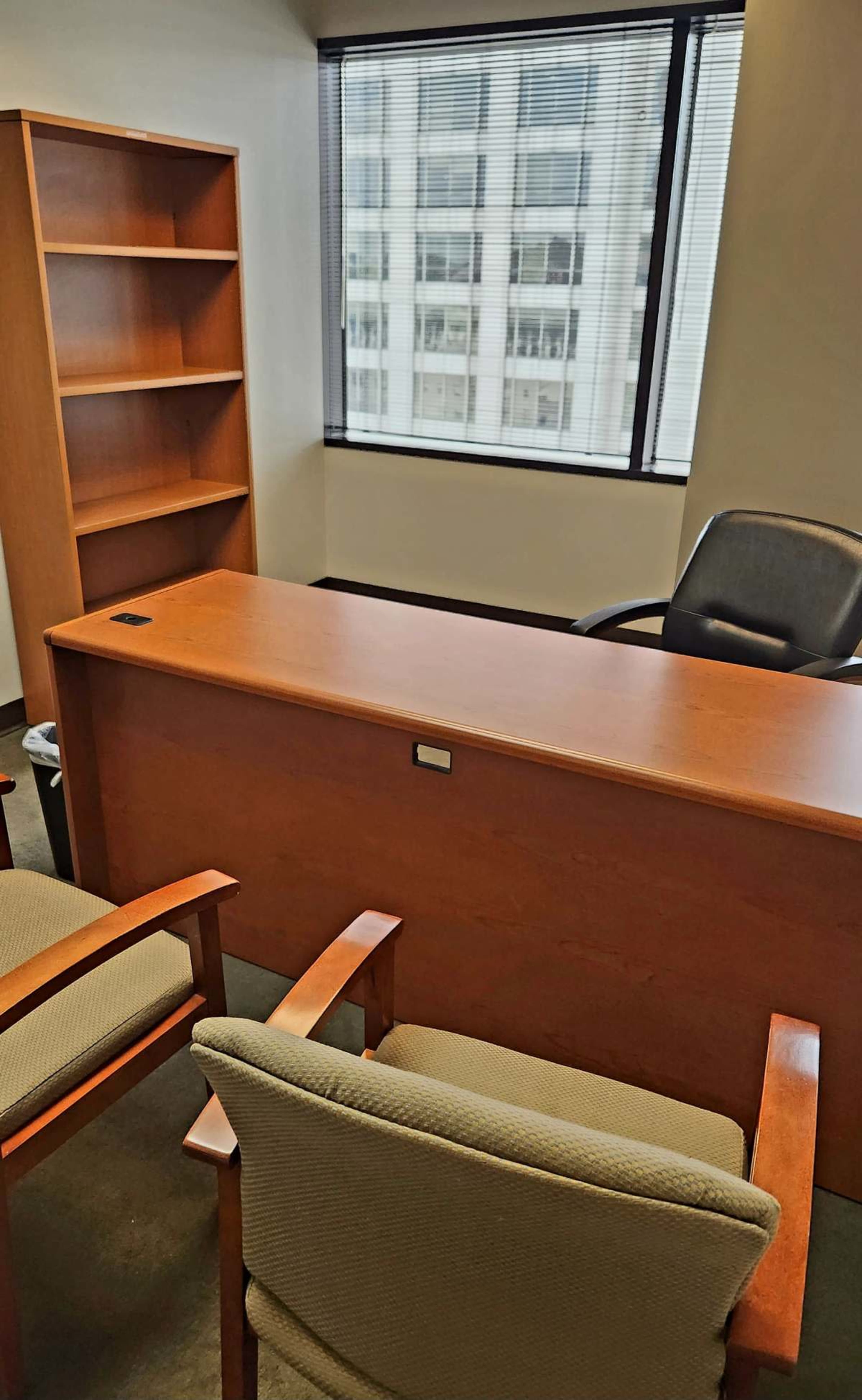 An office space featuring a wooden desk, two chairs, and a bookshelf, with large windows revealing a view of a cityscape.
