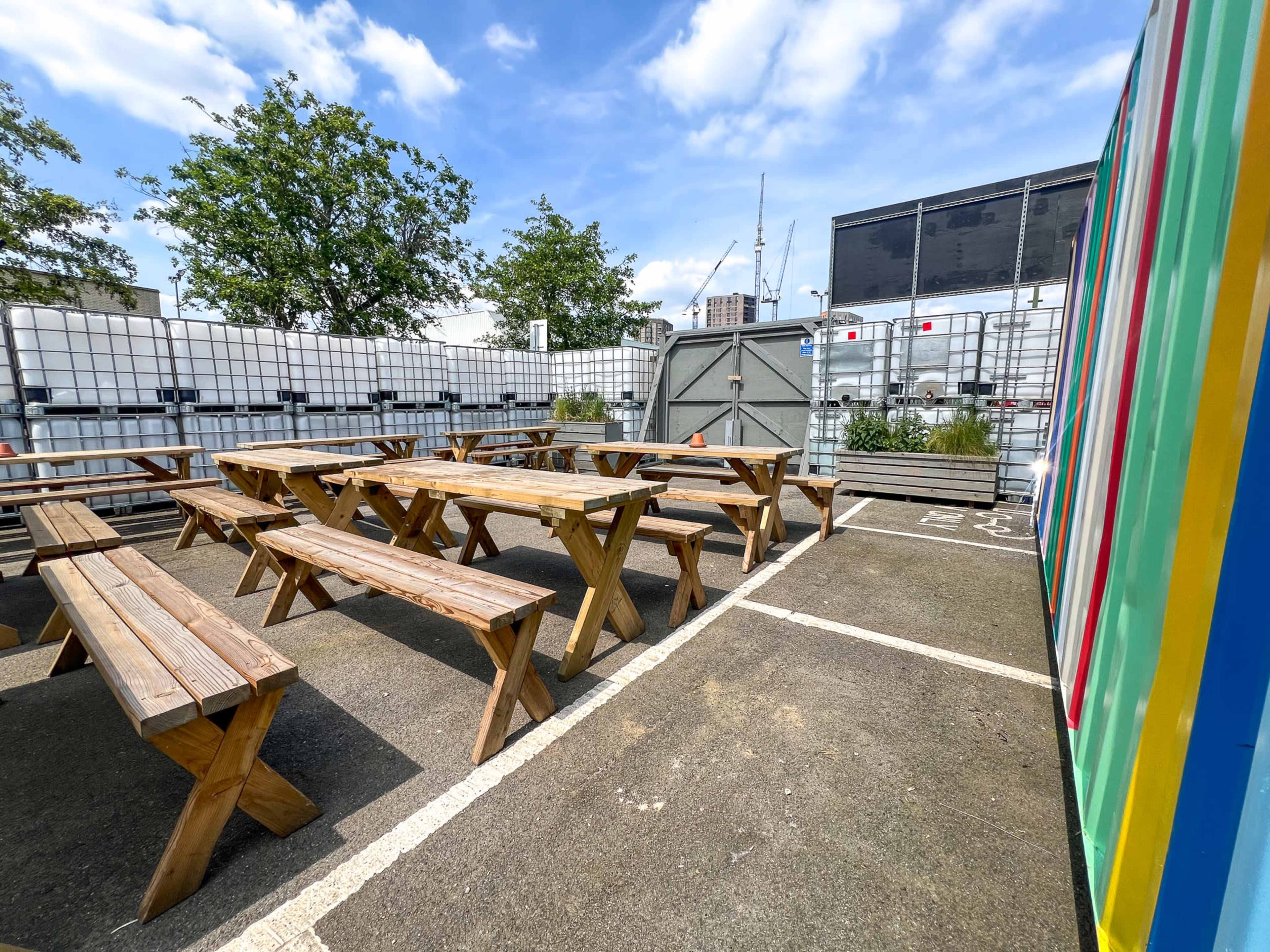 An outdoor seating area with wooden picnic tables surrounded by industrial containers and greenery under a blue sky.
