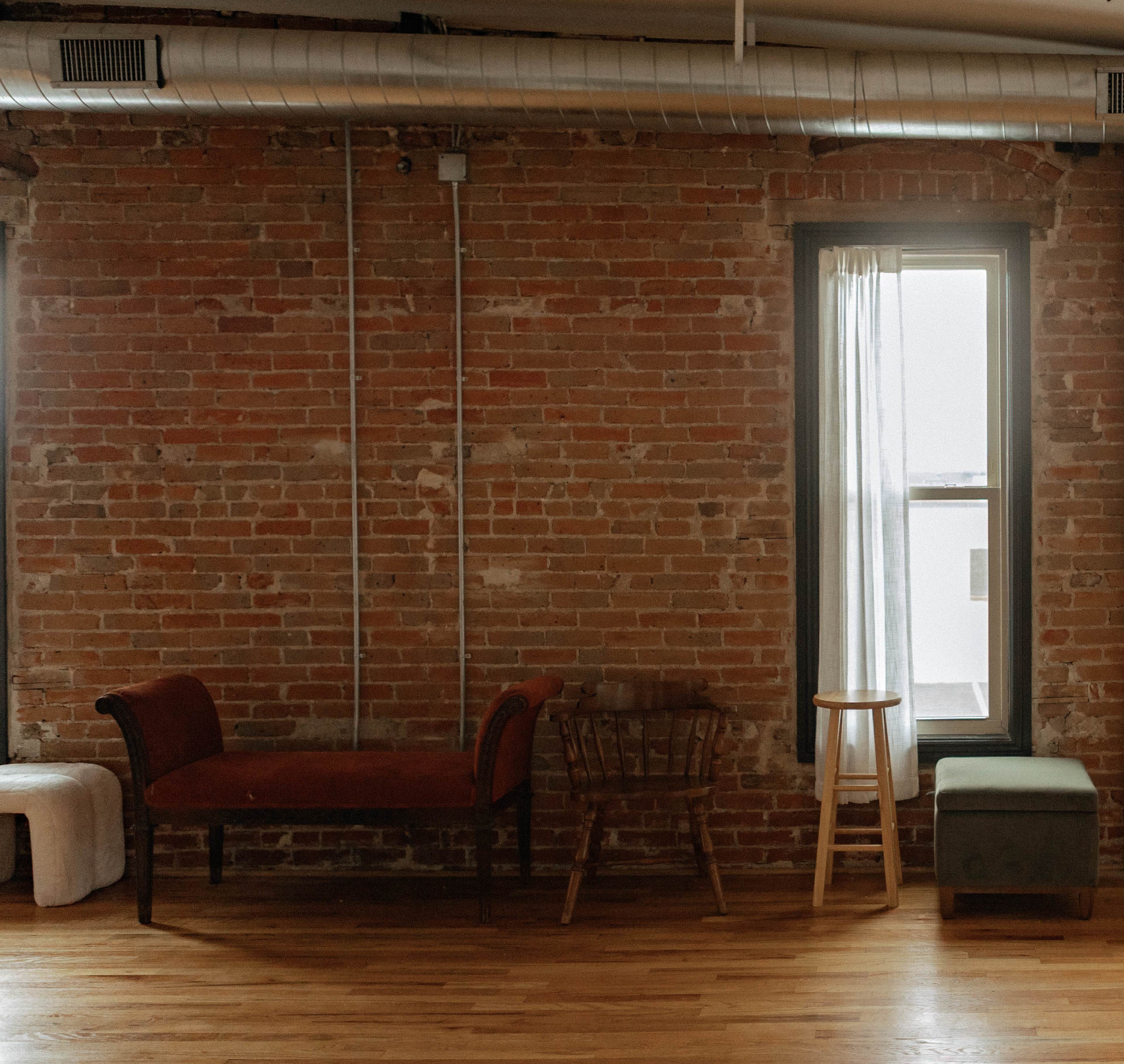 The image shows a room with exposed brick walls, a window draped with a sheer curtain, and three pieces of furniture: a red sofa, a wooden chair, and a short stool.
