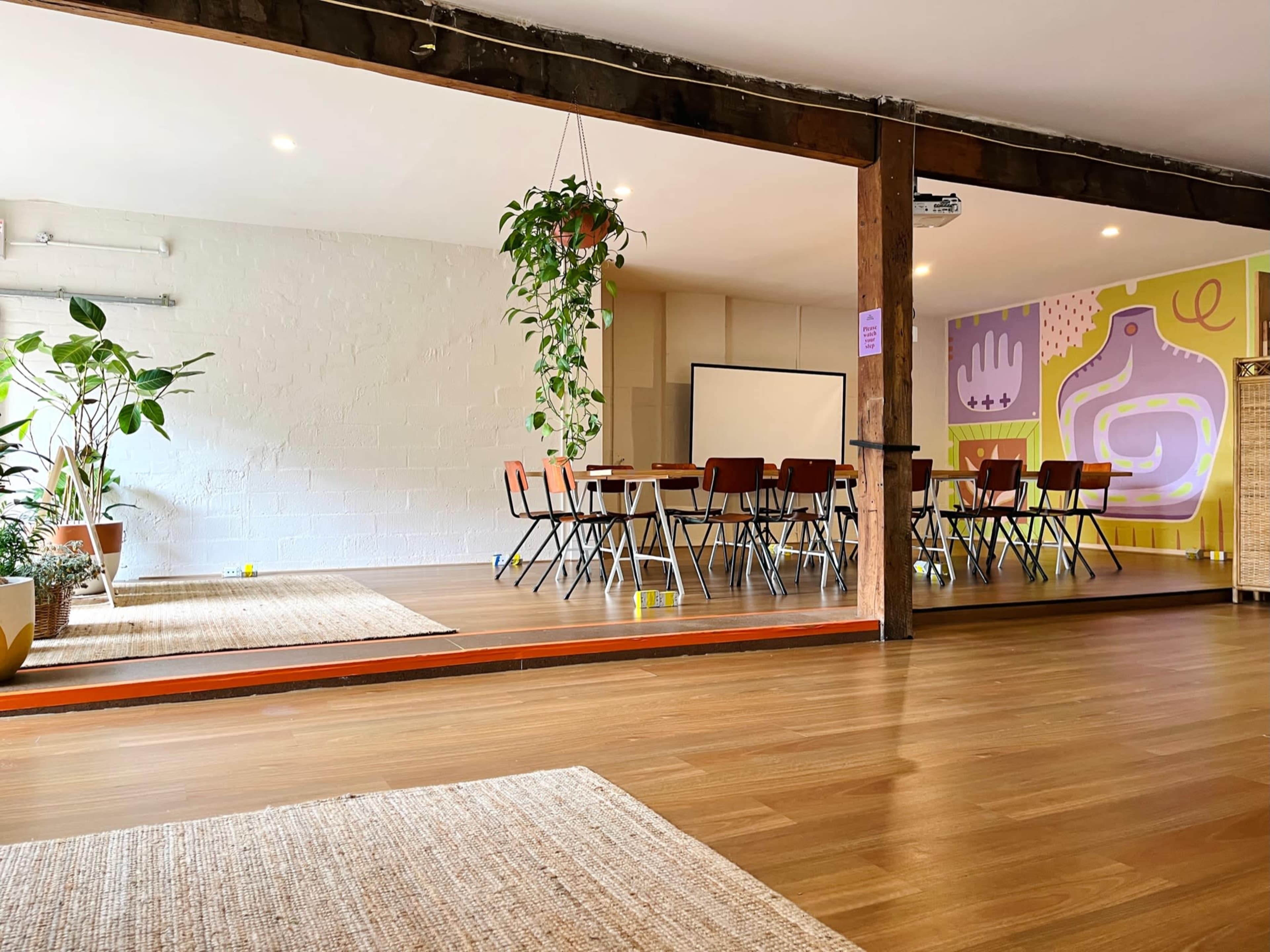 The image shows a spacious room with wooden floors, featuring a long table surrounded by chairs and decorated with plants and large wall murals.