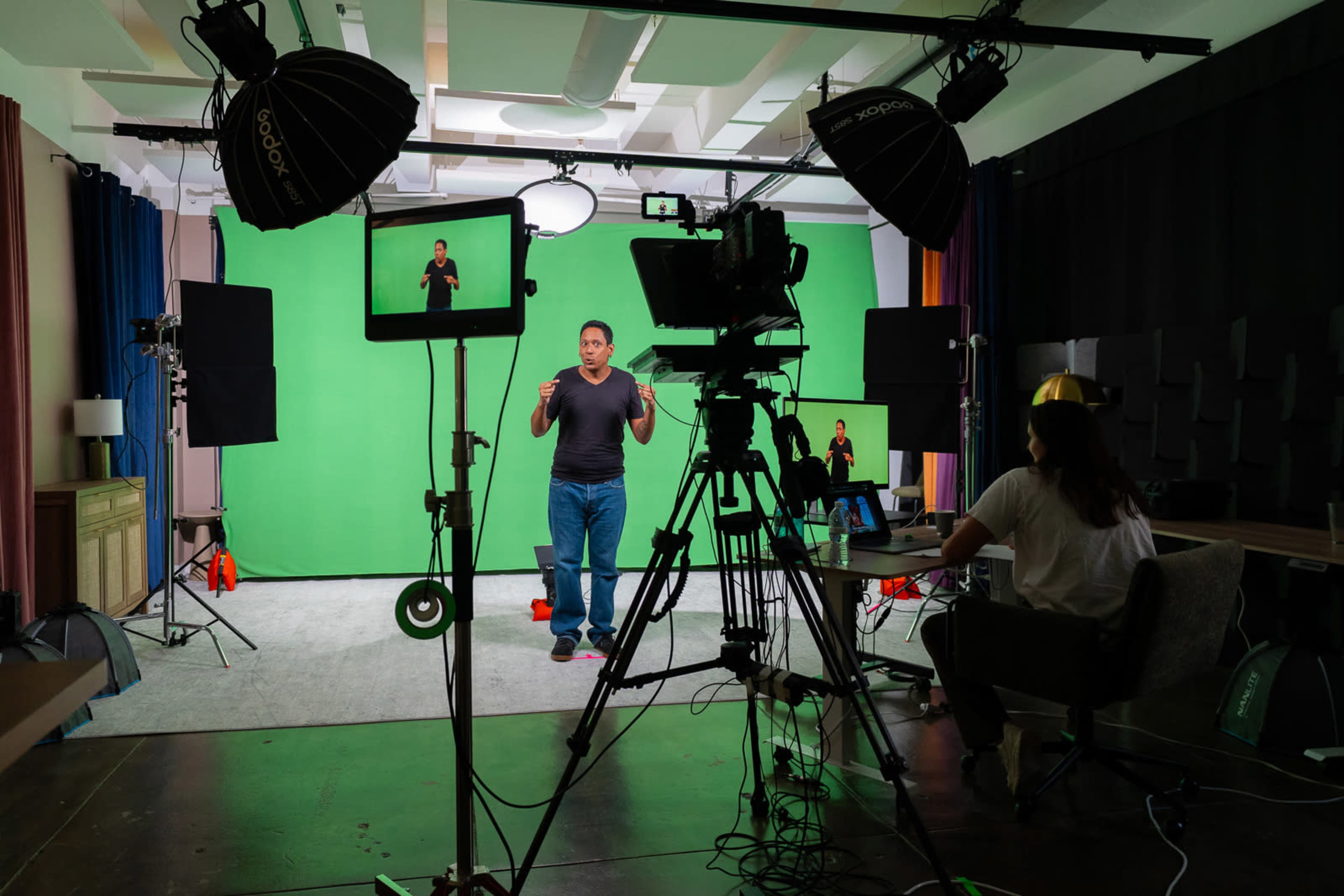 A studio setup features a person speaking in front of a green screen, surrounded by cameras, lighting equipment, and a seated crew member at a desk.