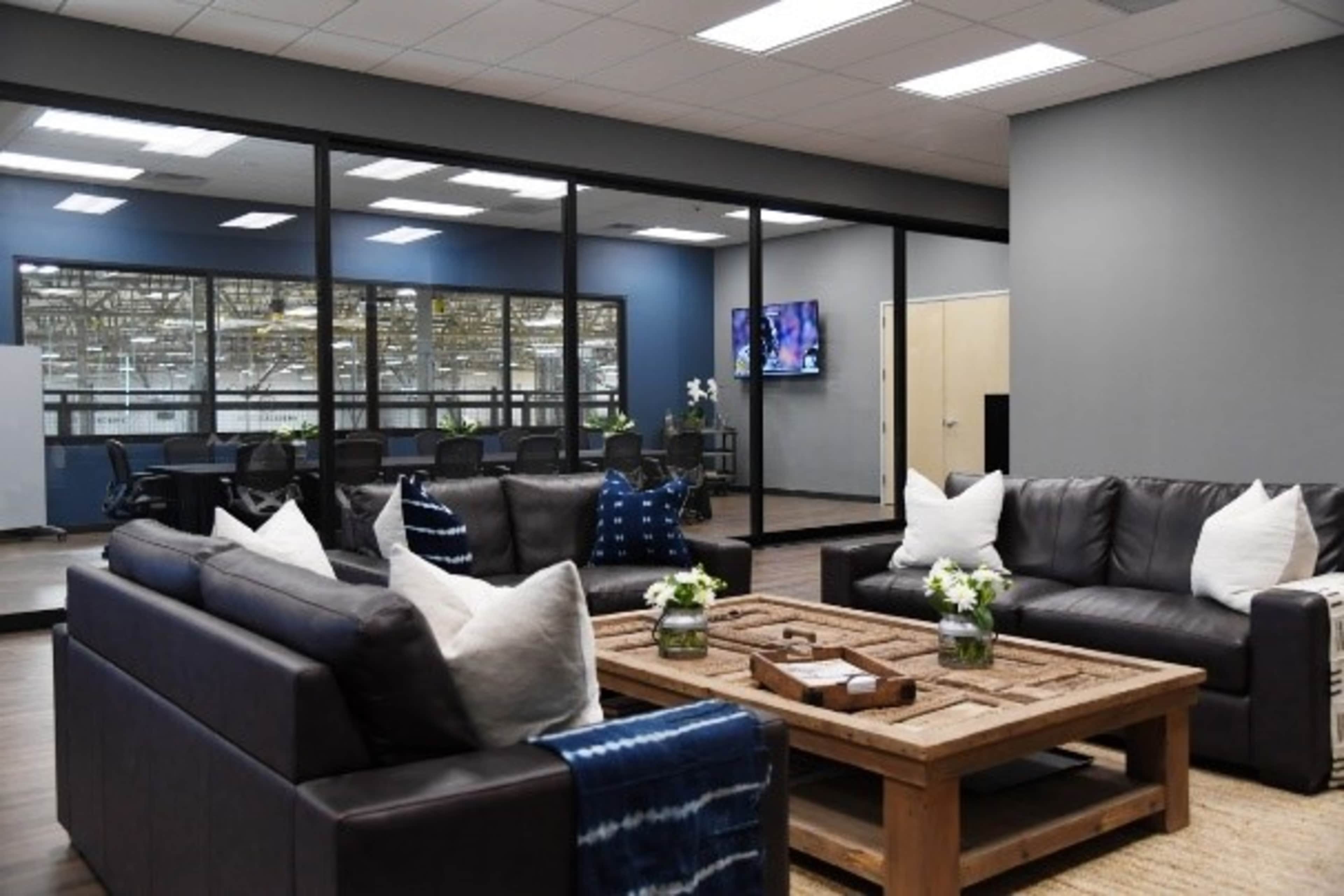 A modern office lounge area with dark leather sofas, a wooden coffee table, and a glass-walled conference room in the background.