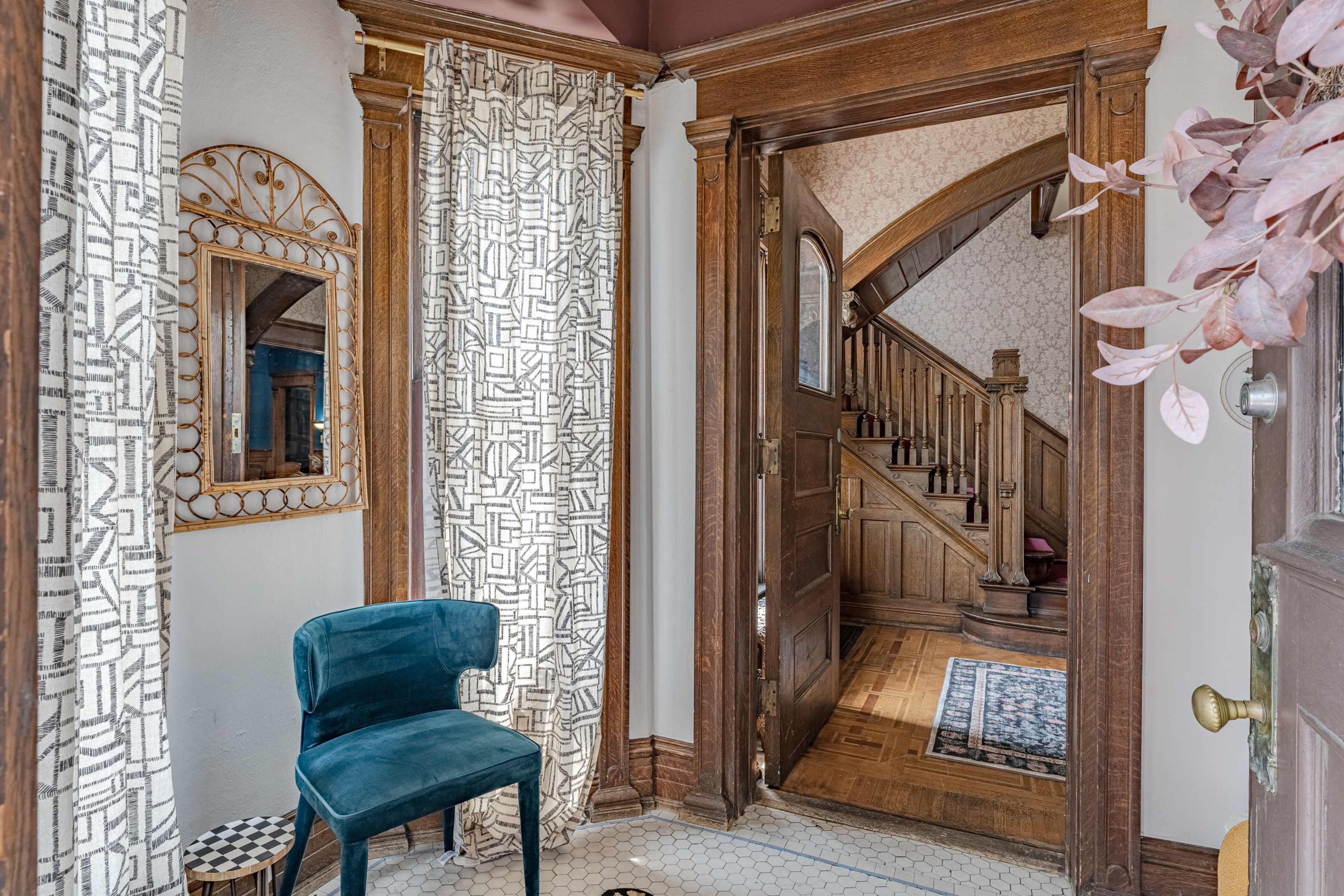 A hallway entrance with a staircase visible in the background, adorned with patterned curtains, a mirror, and a blue chair.
