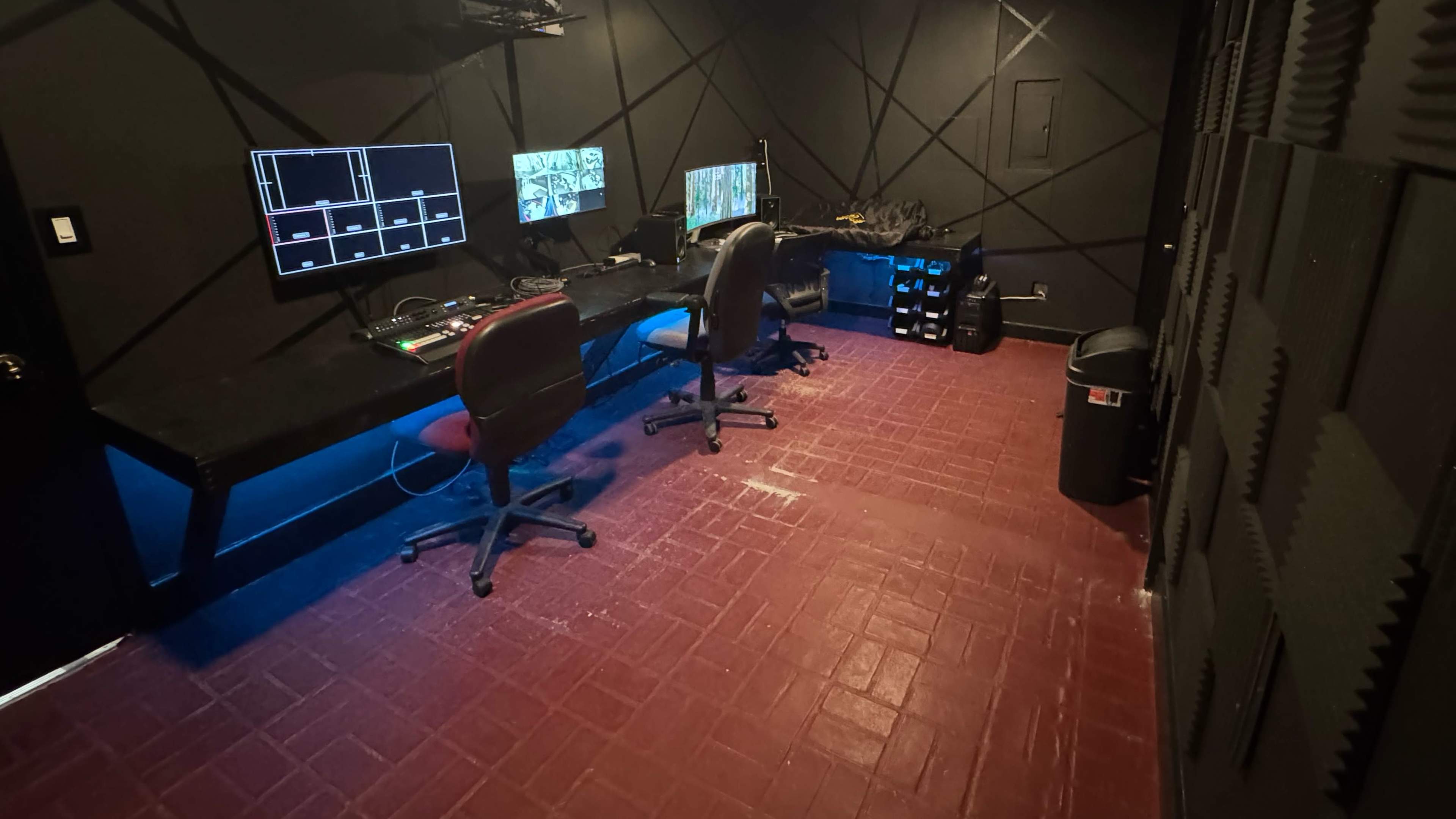The image shows a dimly lit control room with two office chairs, multiple computer monitors on a long desk, and soundproofing panels on the walls.