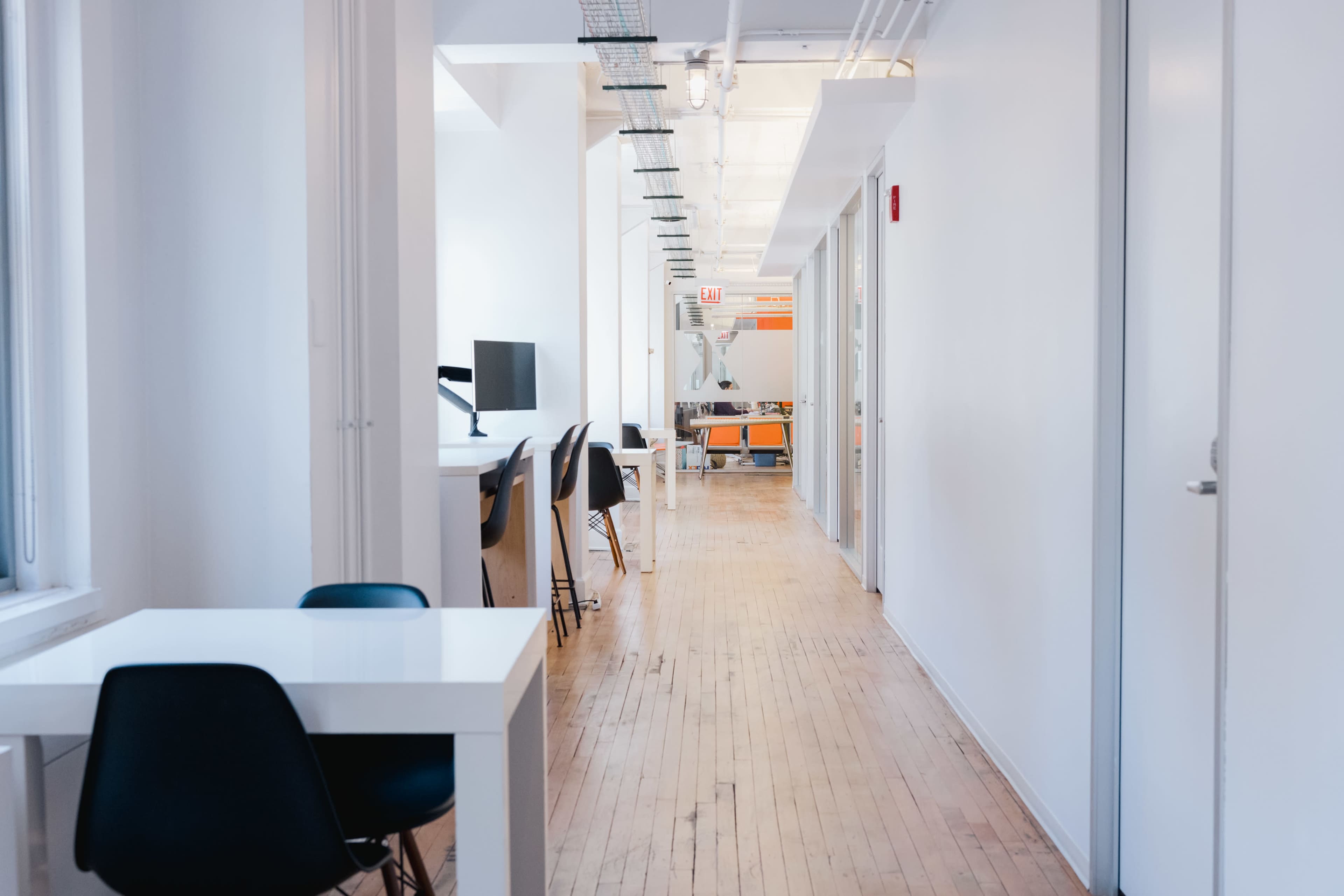 The image shows a clean, bright office hallway with a series of desks on one side and closed doors along both sides.