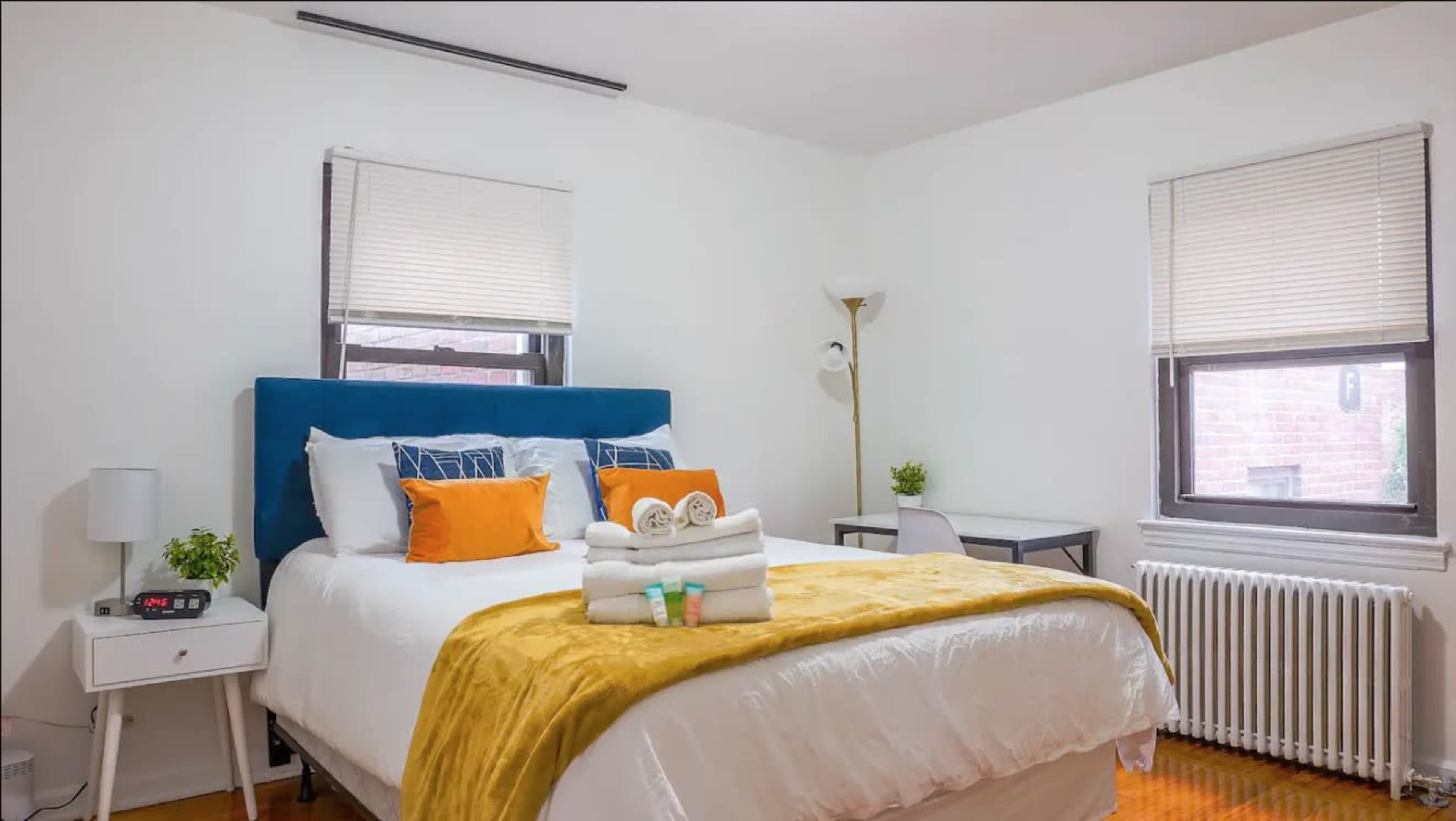 A neatly arranged bedroom features a blue upholstered headboard, a white bed with colorful cushions, and a small desk near a window.