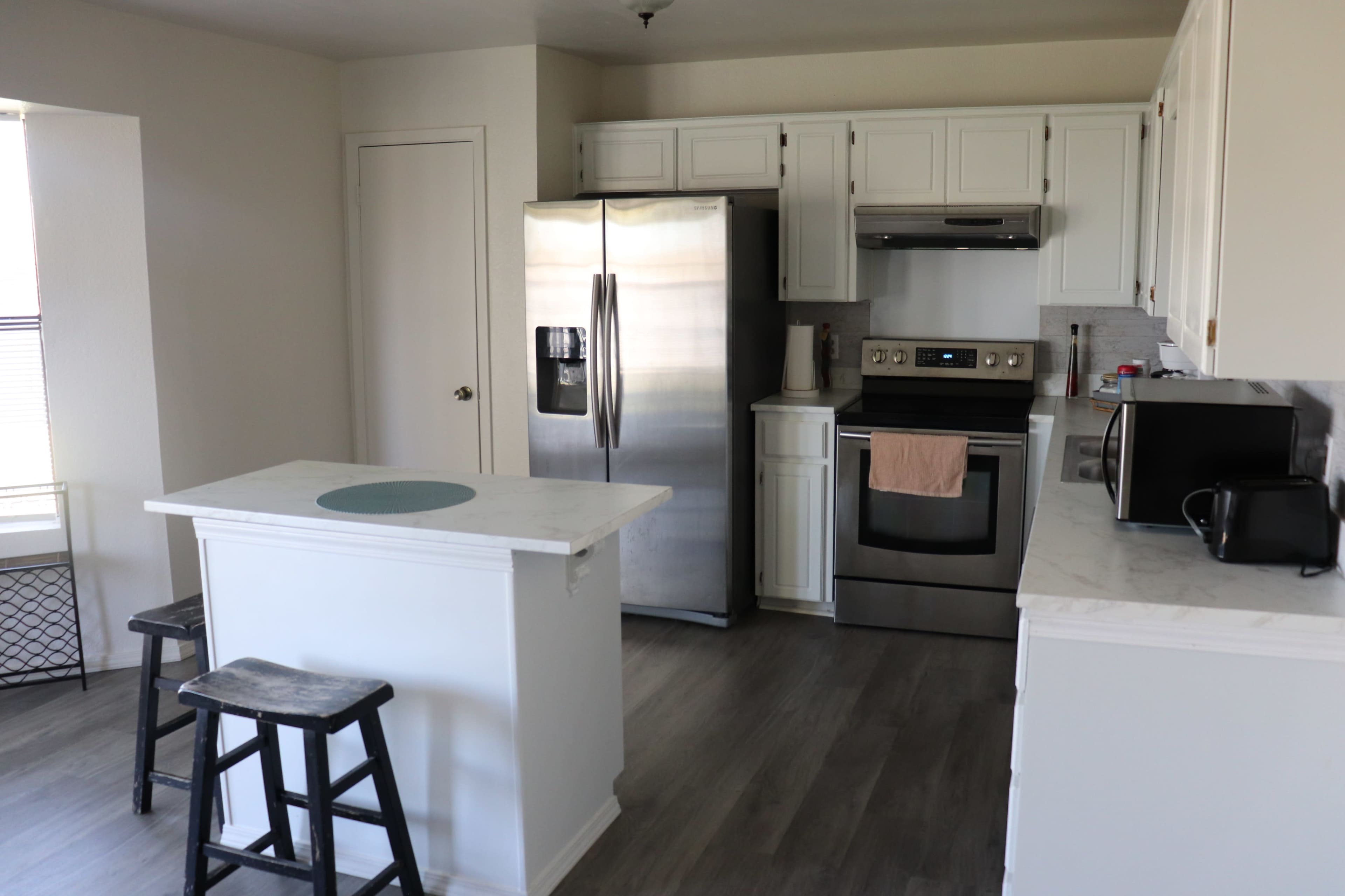 A modern kitchen features stainless steel appliances, white cabinetry, and minimal furnishings including a small table and stools.